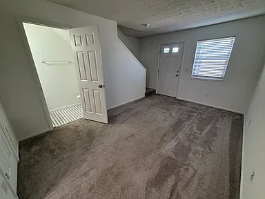 Empty living room with beige carpet, white walls, a window with blinds, a front door, and an open closet with a white door.
