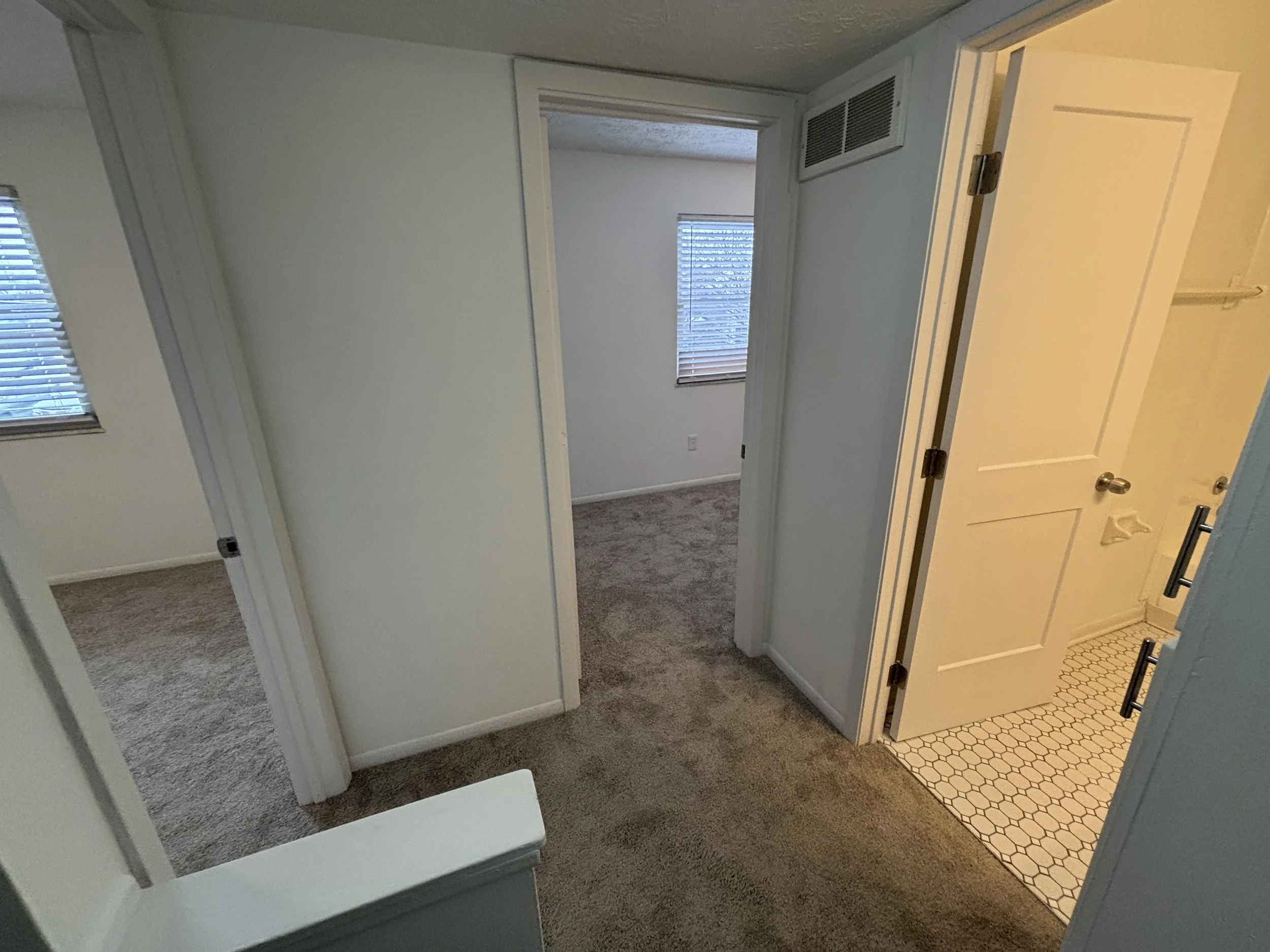 Interior view of a home with multiple rooms, including a bedroom with windows and blinds, and a bathroom with tiled floor, open door, and visible shower towel bar.