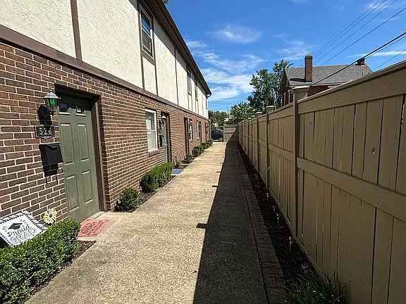 A narrow side walkway running alongside a brick residential building with a green door, a mailbox, potted plants, and a wooden fence on the opposite side, under a blue sky with some clouds.