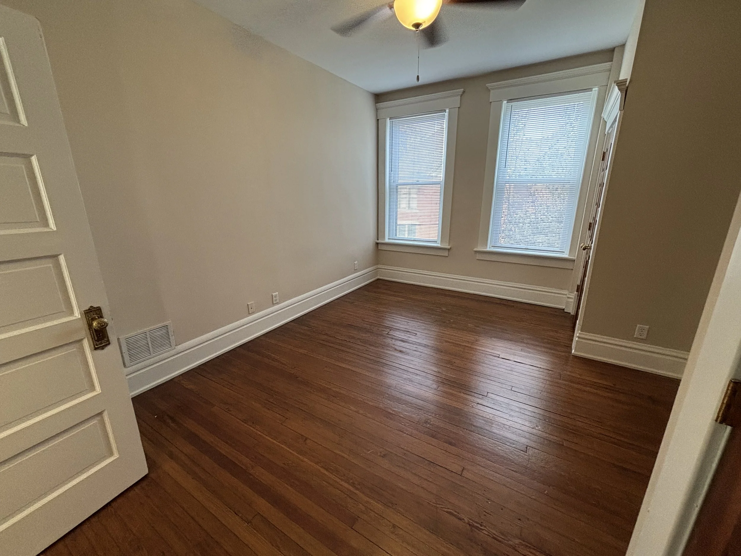 Empty room with hardwood floors, two large windows with blinds, white walls, ceiling fan with light, and a closed door.