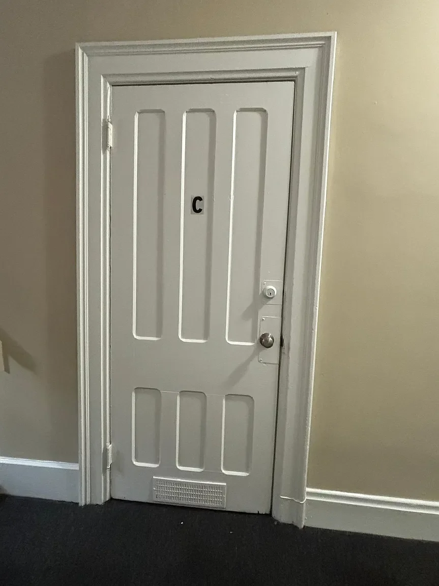 White door labeled with a black letter C, with a doorknob, a lock, and a small vent at the bottom, within a beige wall and dark carpet.