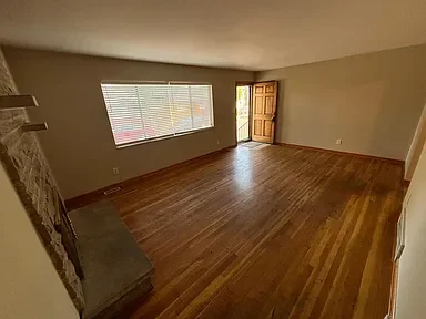 Empty living room with hardwood floors, large window, and a wooden front door.