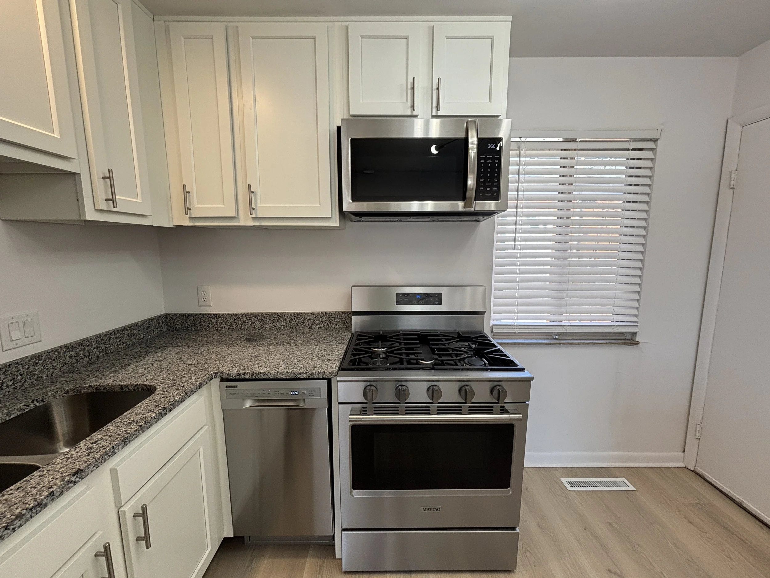 Kitchen with white cabinets, granite countertops, stainless steel appliances including a microwave, stove, and dishwasher, window with blinds, light wood flooring, and a door.