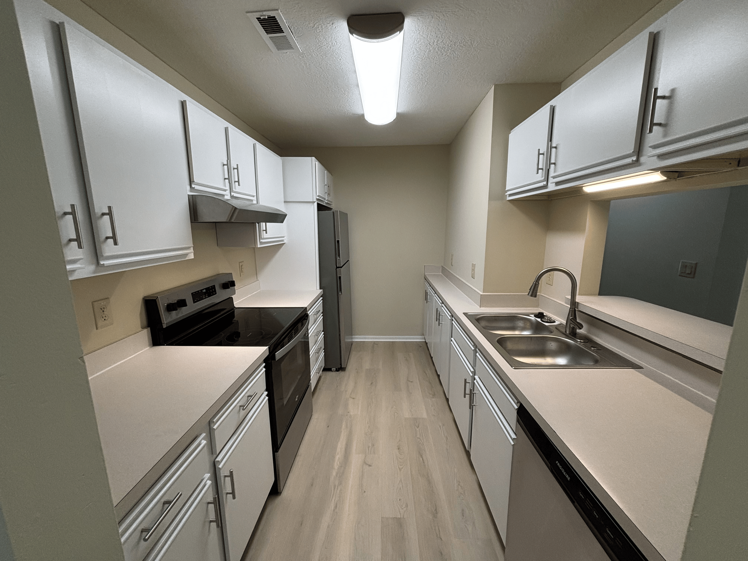 Empty kitchen with white cabinets, black stove, refrigerator, and dual sink, beige walls, light wood flooring.