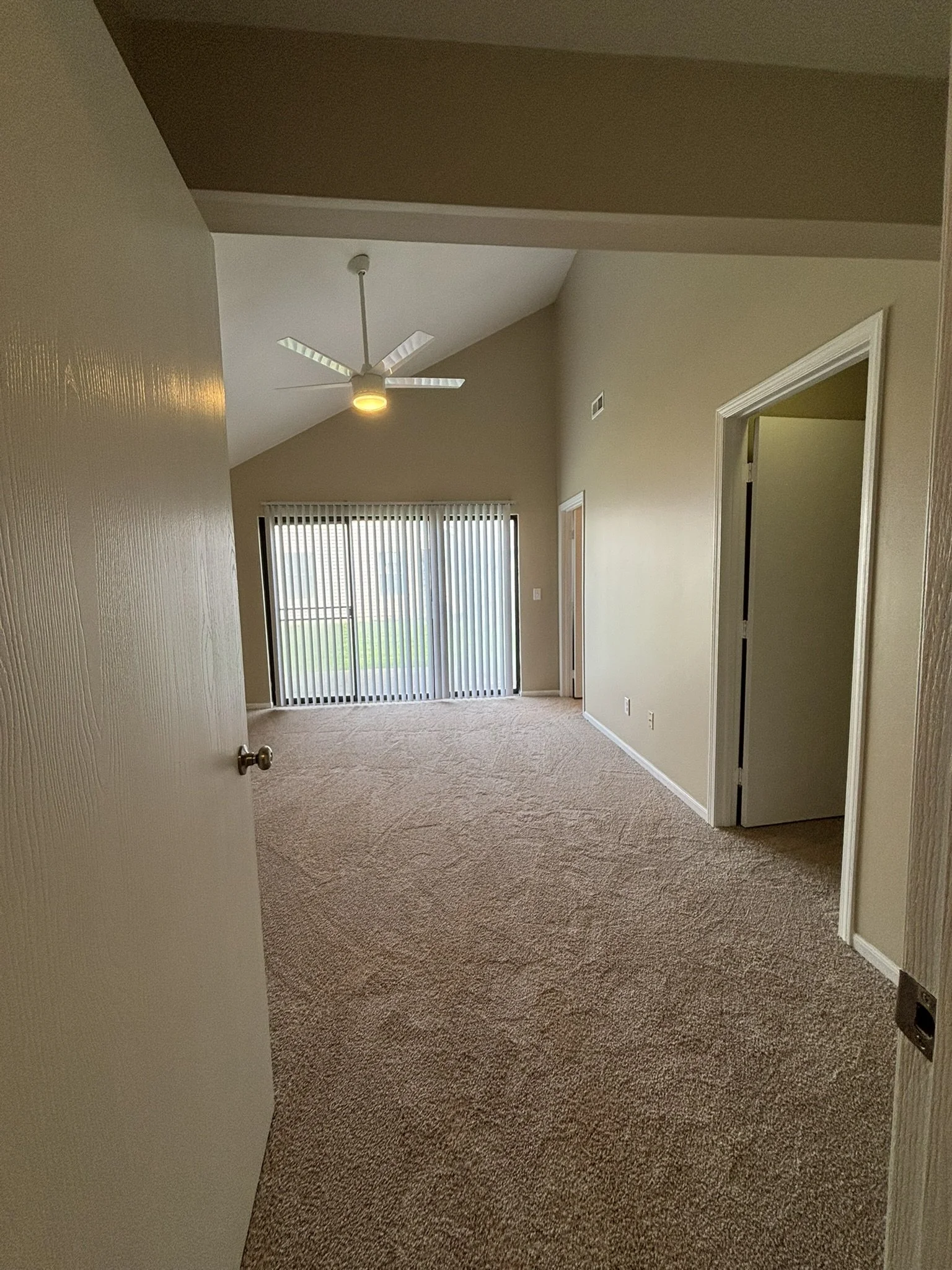 Empty living room with beige carpeting, a ceiling fan, sliding glass door with vertical blinds, and beige walls.
