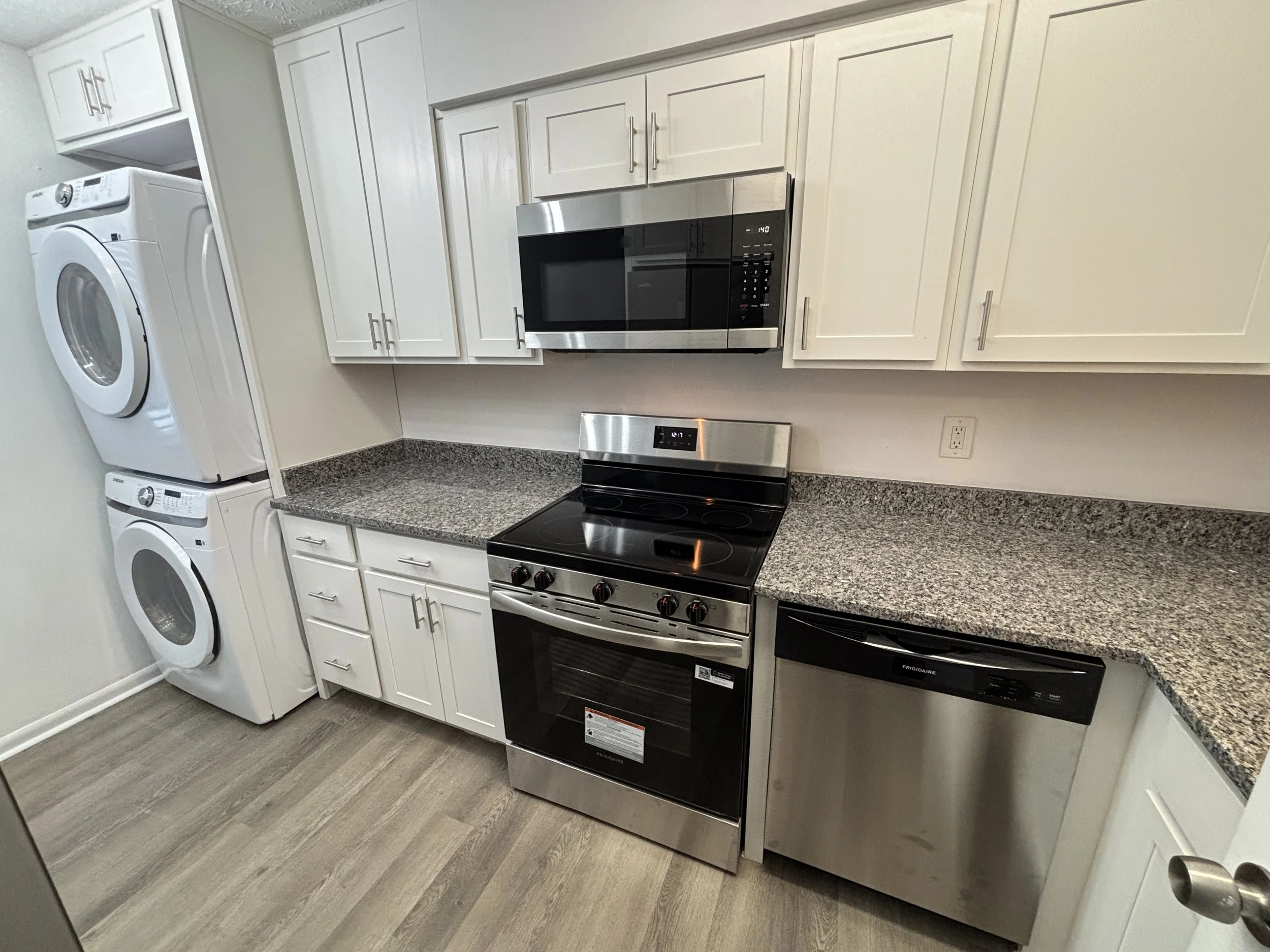 Kitchen with white cabinets, granite countertop, stainless steel appliances including microwave, oven, and dishwasher, and a stacked washer and dryer.