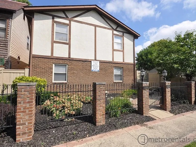 A two-story house with brick and white paneled exterior, surrounded by a black metal fence and landscaped garden bed, on a bright, sunny day.