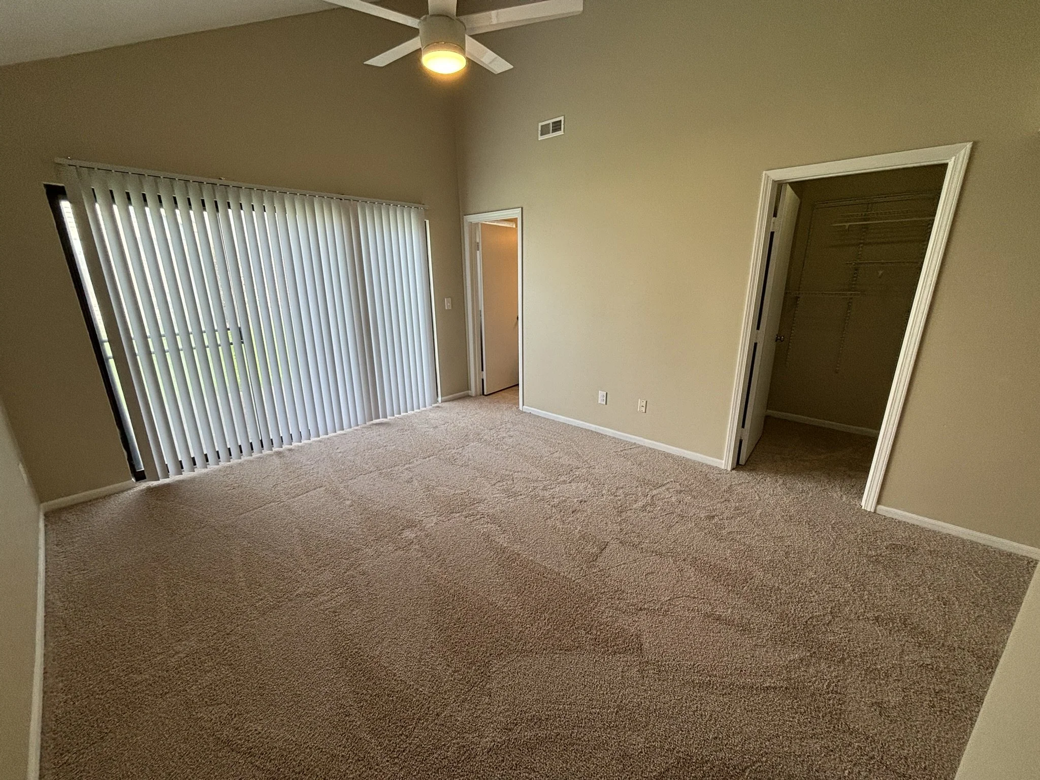 Empty bedroom with beige carpet, beige walls, a ceiling fan, sliding glass door with vertical blinds, and a walk-in closet.