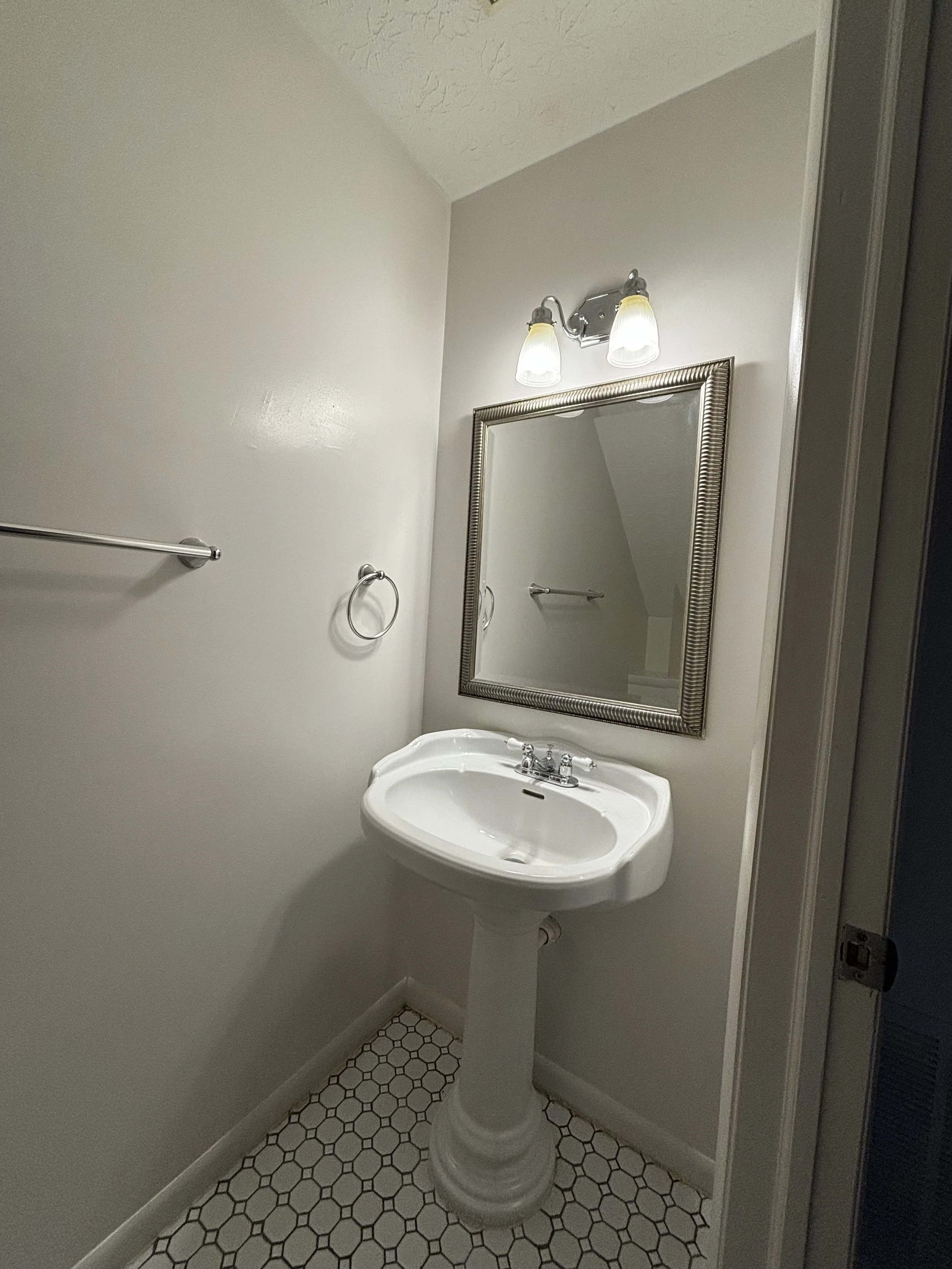 A small bathroom with a pedestal sink and a rectangular mirror above it. There is a light fixture with two bulbs above the mirror, a towel ring to the left of the mirror, and a towel bar on the left wall. The floor has small hexagon white tiles with 