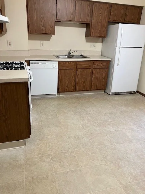 Kitchen with wooden cabinets, a white refrigerator, a dishwasher, a sink, and a beige tiled floor.
