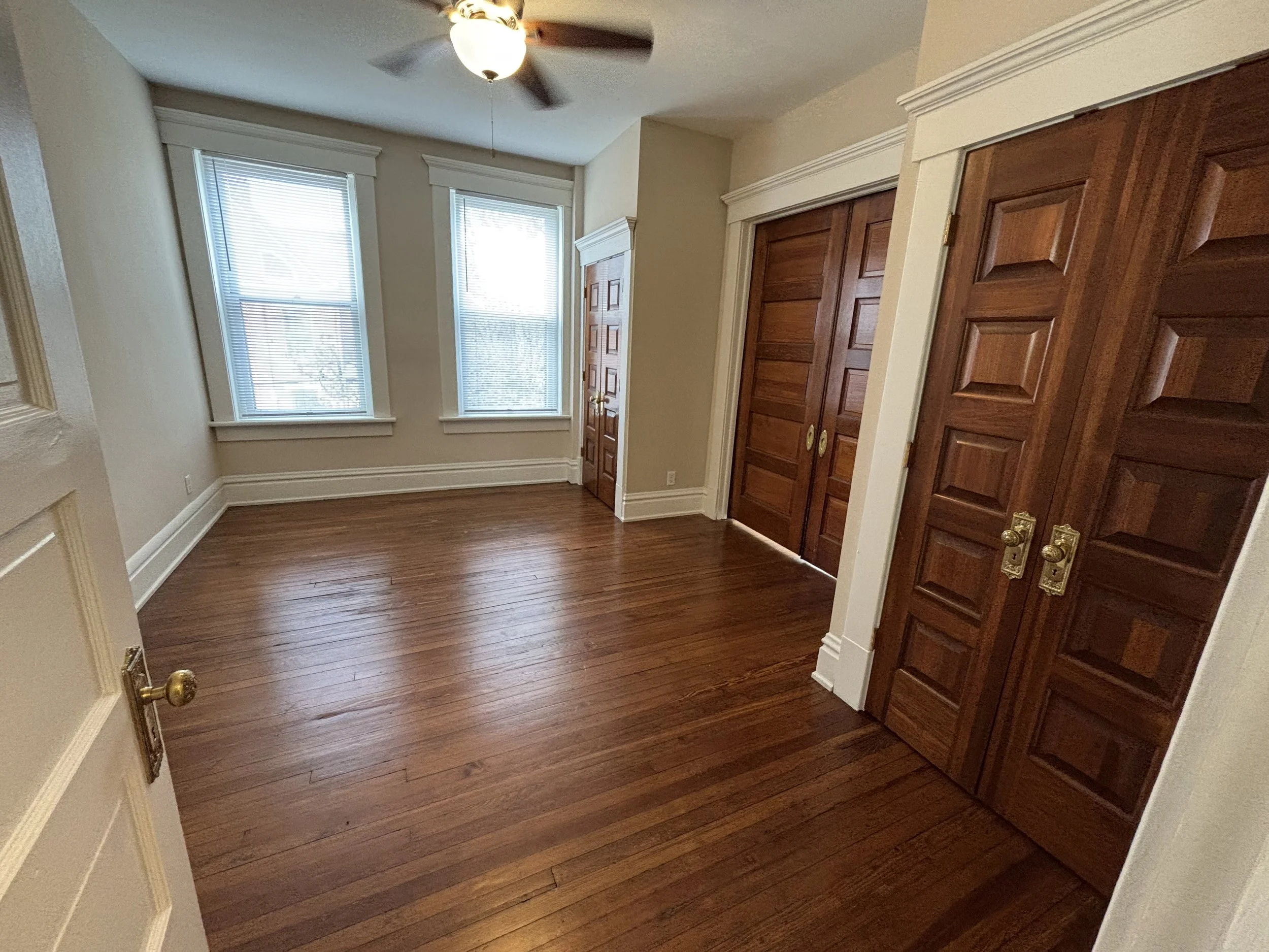 Empty room with hardwood floors, two windows with blinds, a ceiling fan, and wooden closet doors.