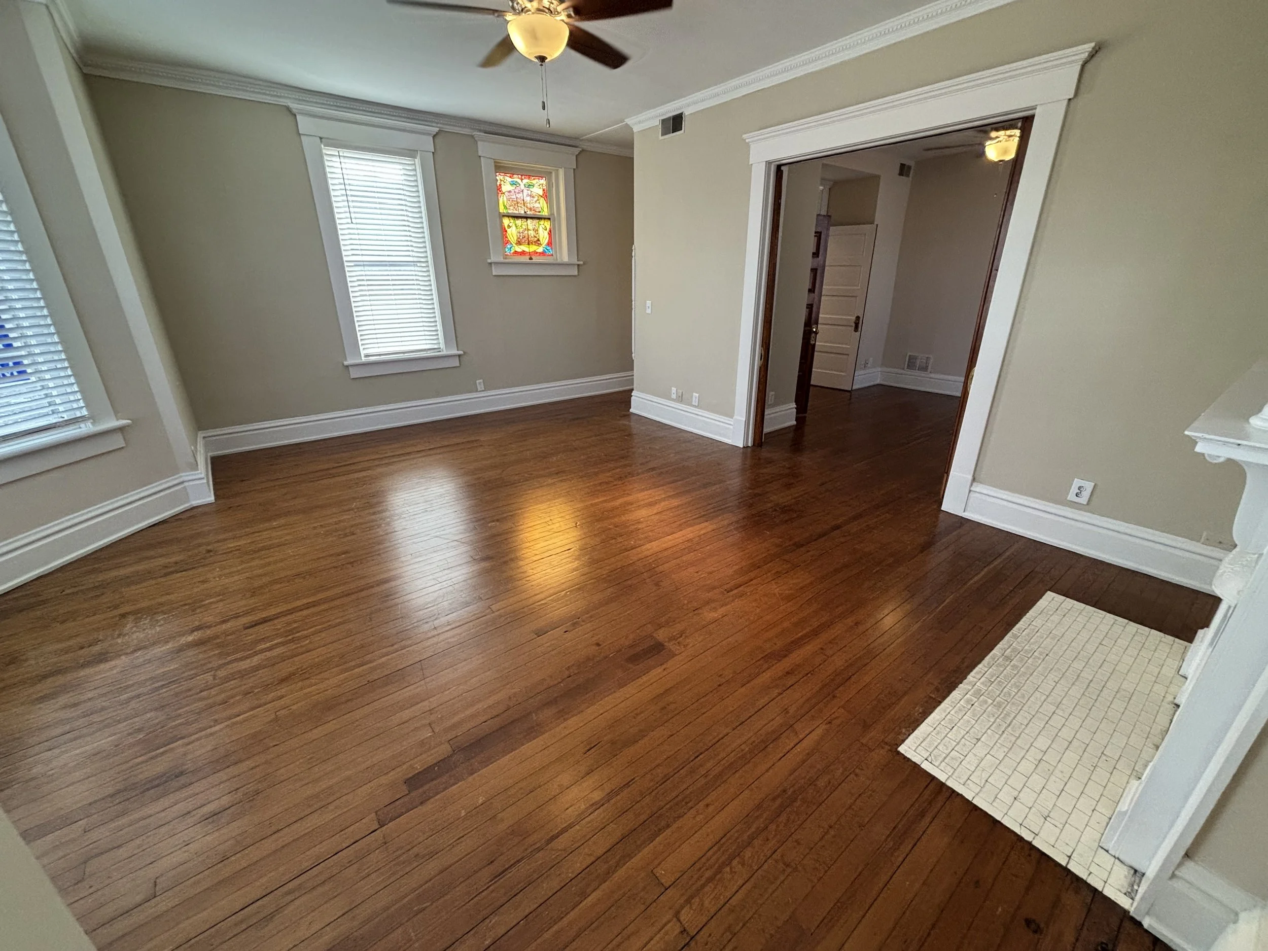 Empty living room with hardwood floors, beige walls, two windows with blinds, a stained glass window, a ceiling fan, and a doorway leading to another room.