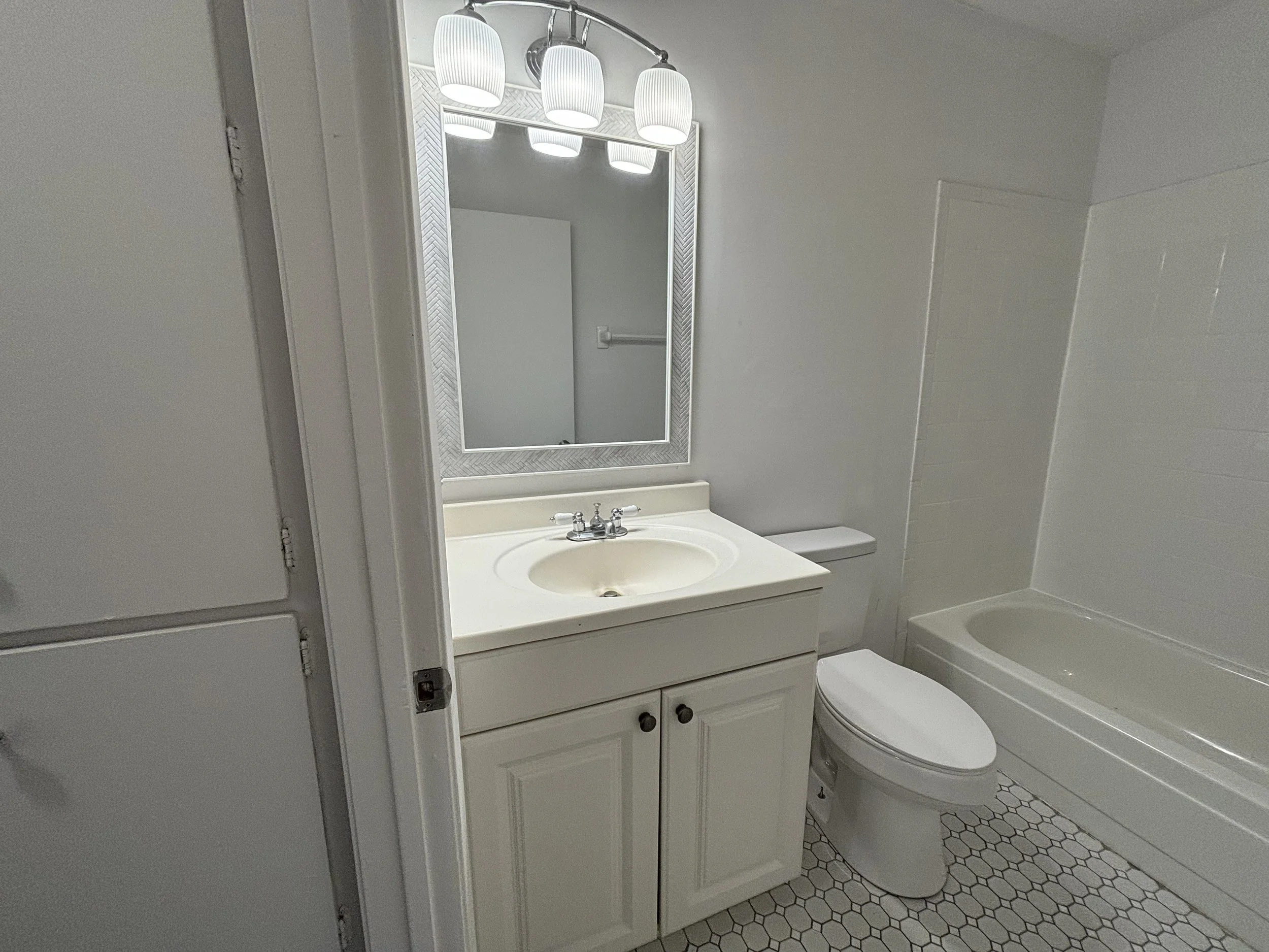Bathroom with white vanity, oval mirror, four light fixtures, toilet, and bathtub with hexagonal tiled floor.