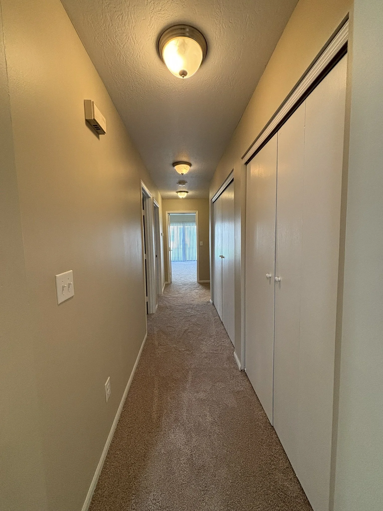 Empty hallway with beige walls, brown carpet, ceiling lights, and closet doors, leading to a room with a sliding glass door.