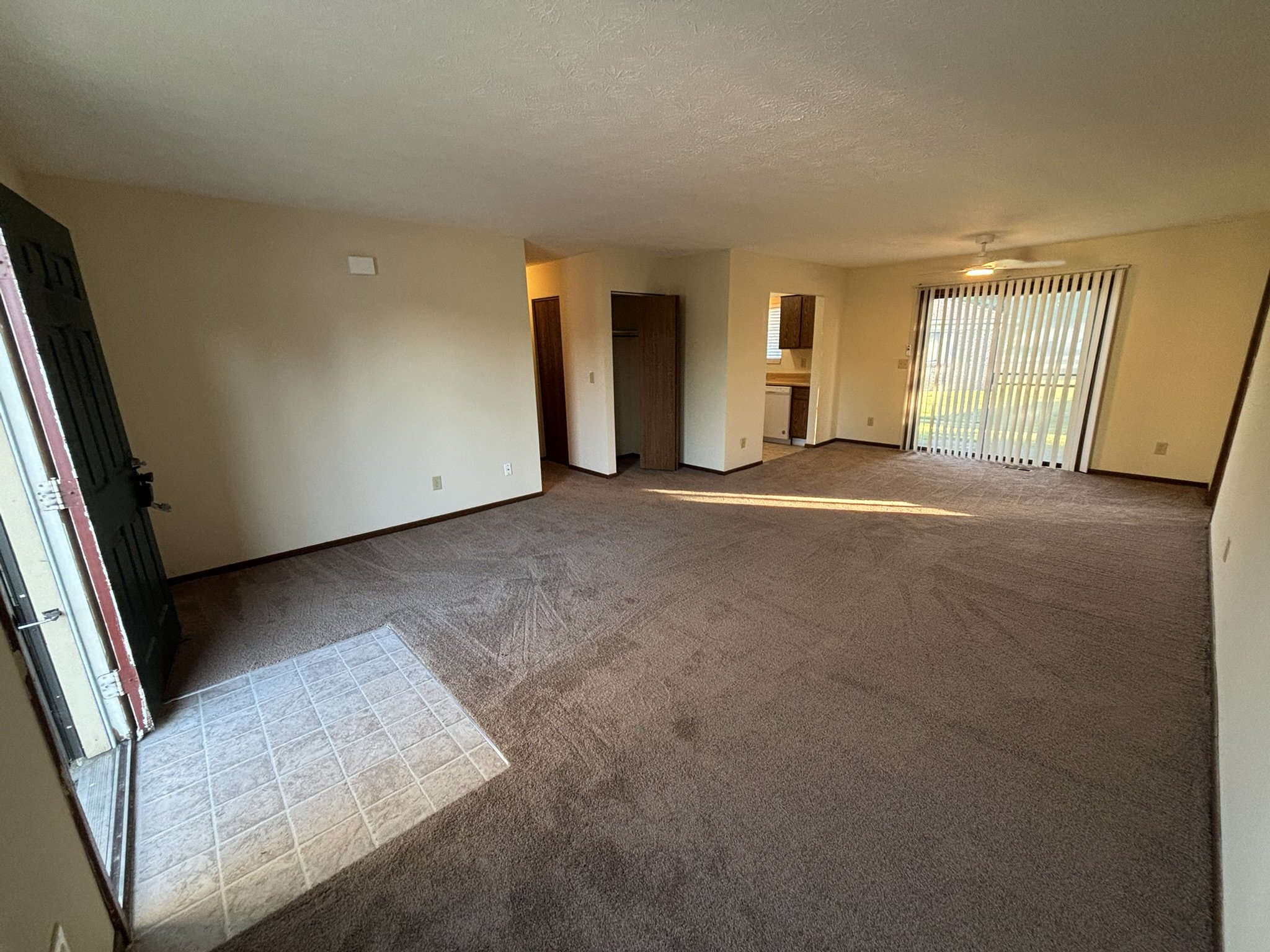 Empty living room with brown carpet, sliding glass door with vertical blinds, small kitchen area, and front door with a tiled entryway.