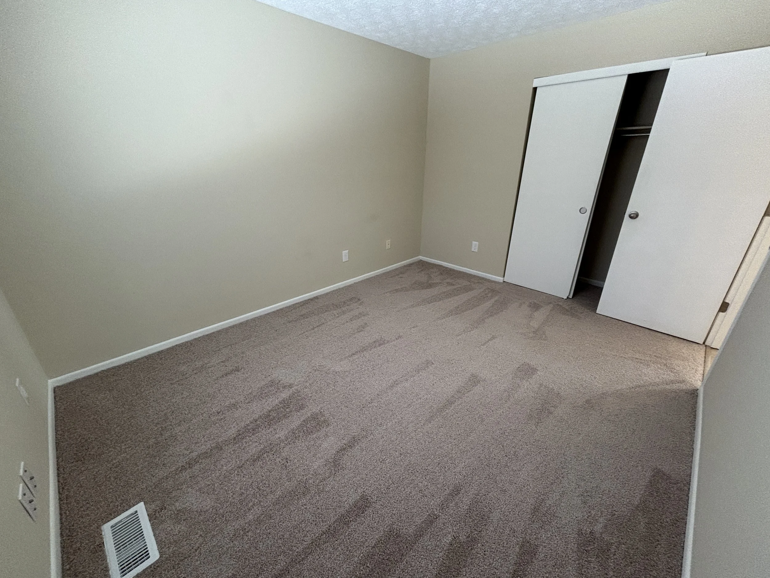 Empty room with beige walls, a beige carpet, a white closet with sliding doors, and a floor vent near the bottom left corner.