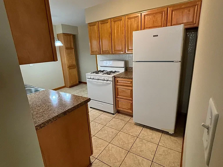 A small kitchen with wooden cabinets, a white refrigerator, a gas stove, a granite countertop, and tile flooring.