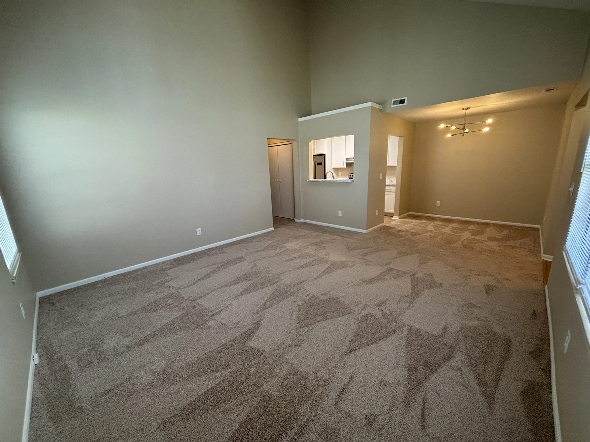 Empty living room with beige walls, carpeted floor, large windows, and open view of the kitchen and dining area.