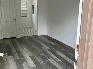 Empty room with dark wooden flooring, white walls, and an open door leading to another area with a striped rug and a window with blinds.