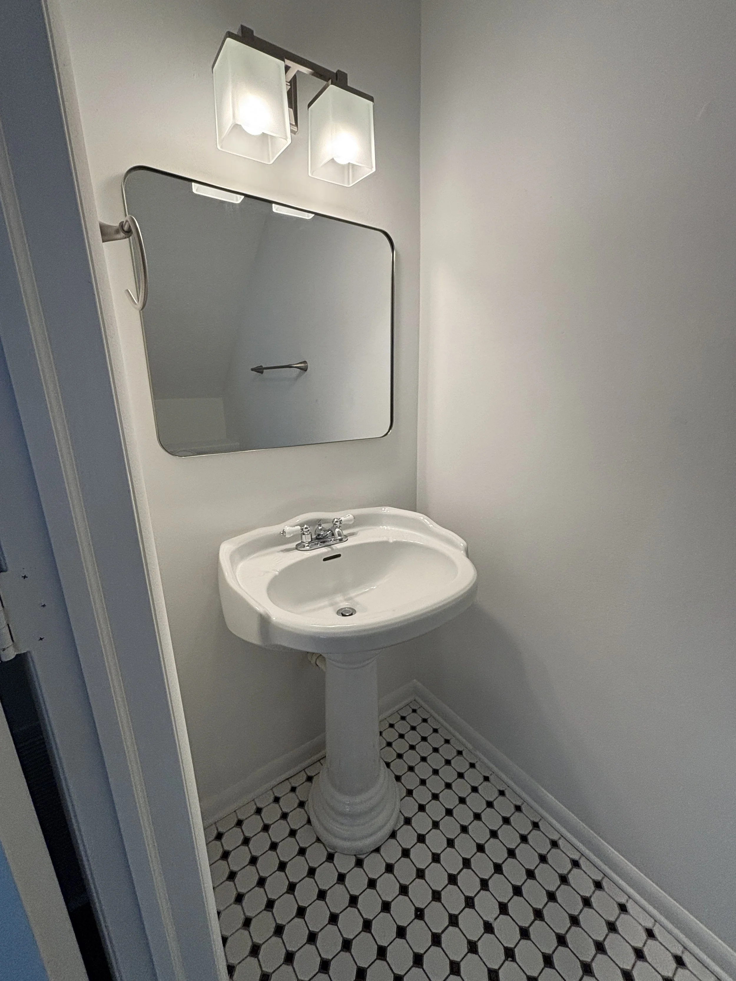 A small bathroom with a pedestal sink, a mirror above it, and a light fixture with two square glass shades mounted above the mirror. The floor has black and white hexagonal tiles, and the walls are painted white.