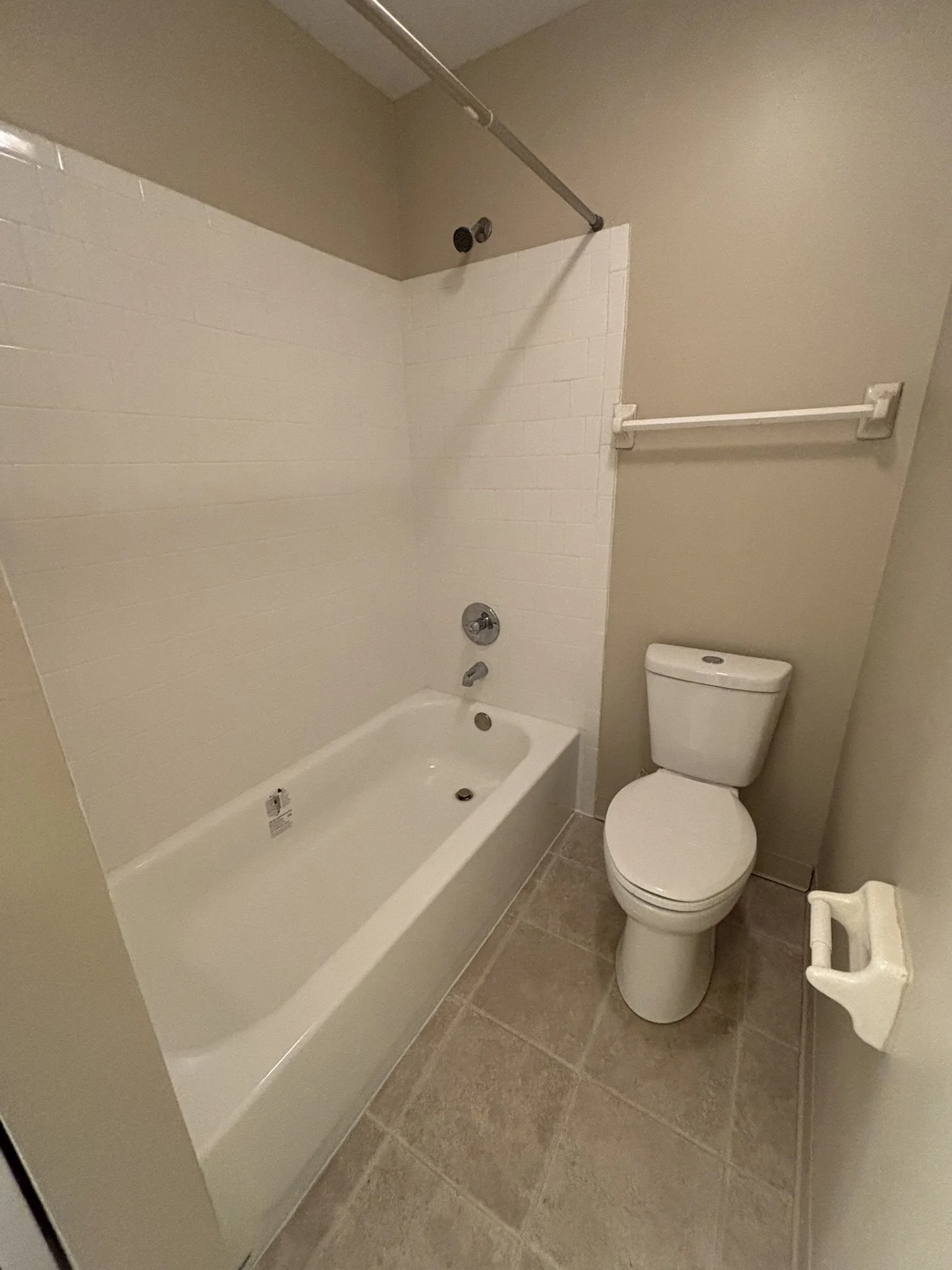 Small bathroom with a bathtub on the left and a toilet on the right, beige tiled floor, white wall tiles in the shower area, beige walls, towel bar above the toilet, and toilet paper holder on the wall.