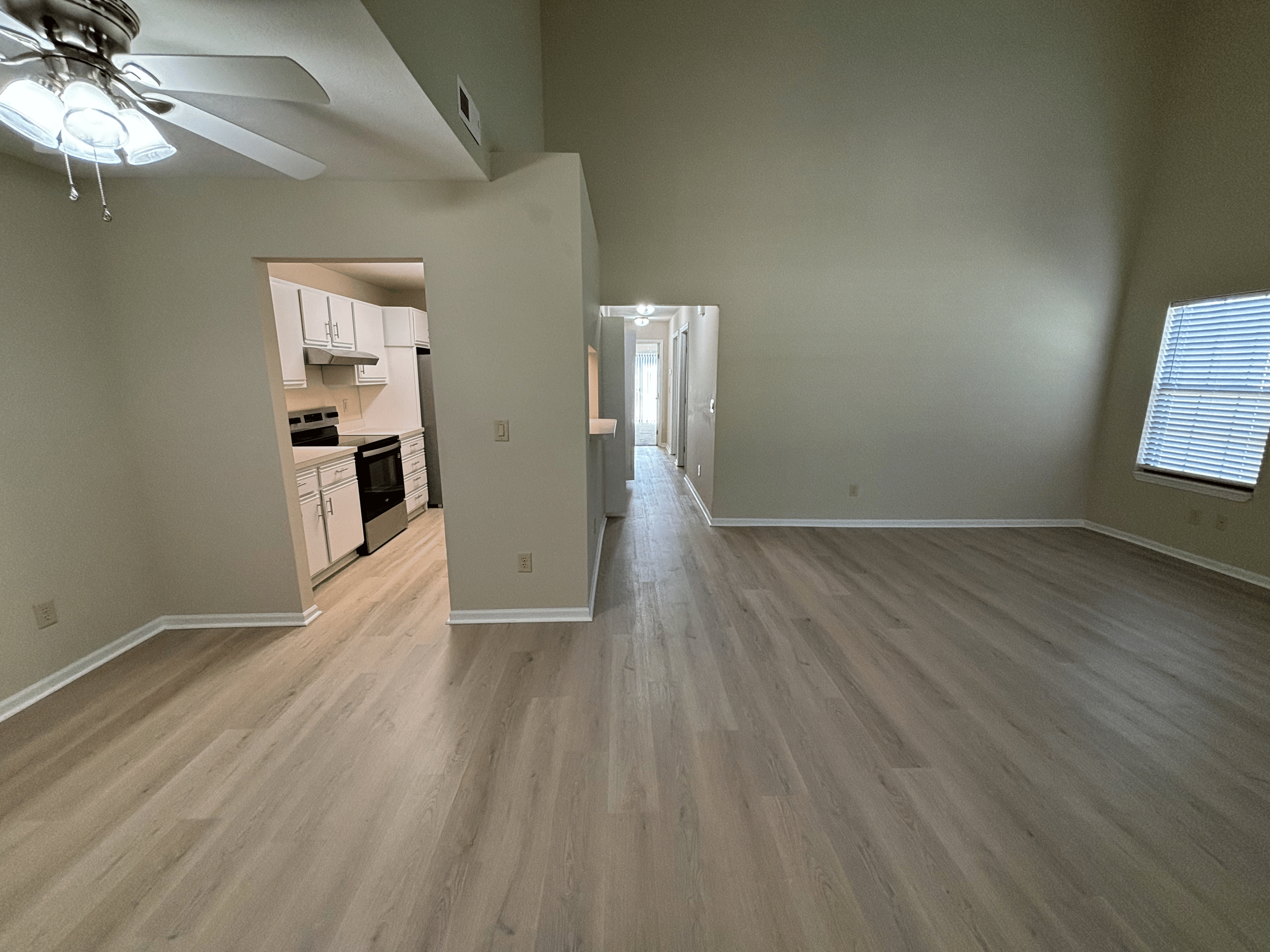 Empty living room with beige walls and wood flooring, windows with blinds, a ceiling fan with lights, and an open kitchen with white cabinets and black appliances in the background.