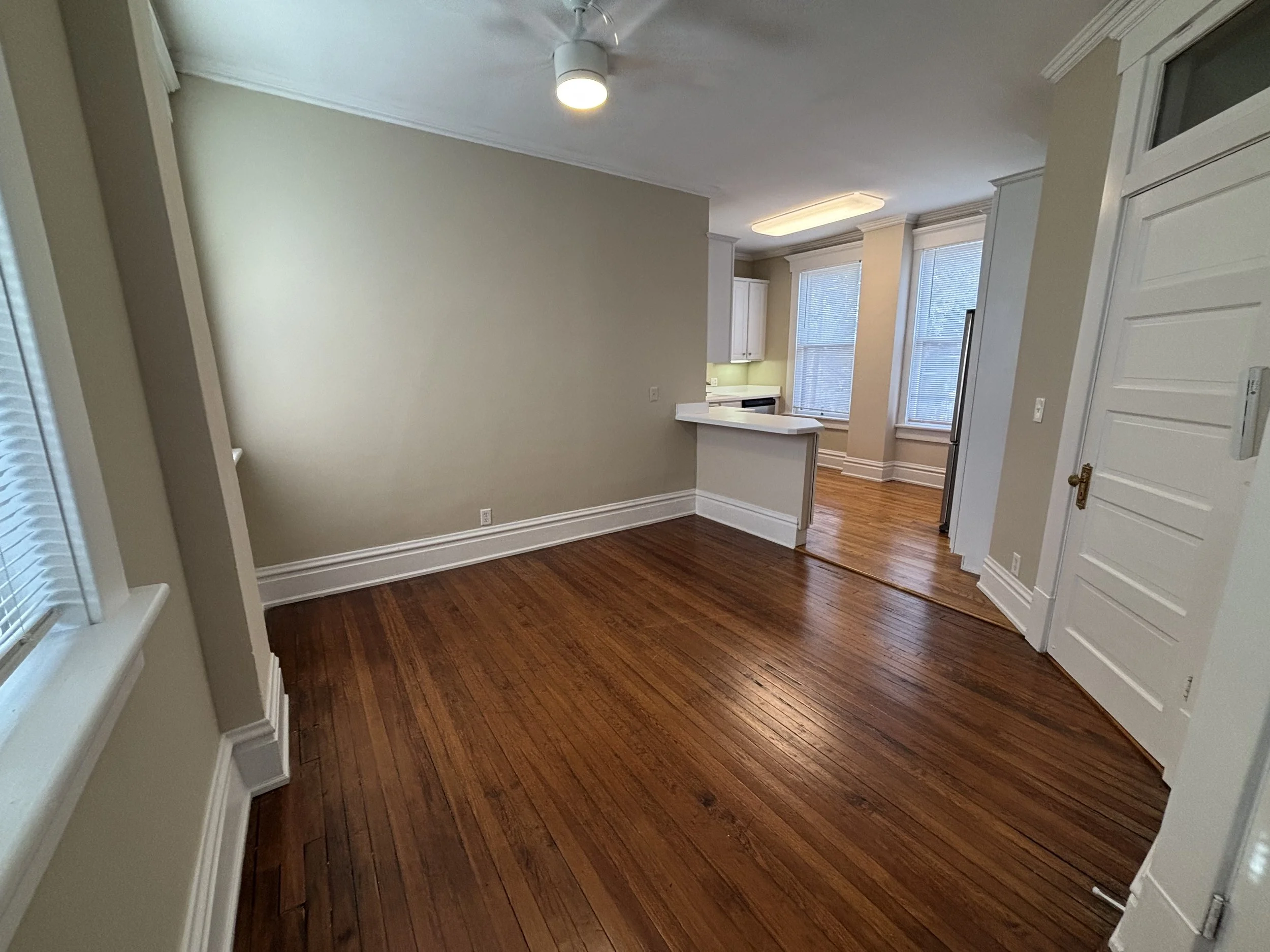Empty living room with wooden floors, light-colored walls, and large windows with blinds, adjacent to a kitchen area with white cabinets and a small breakfast bar.