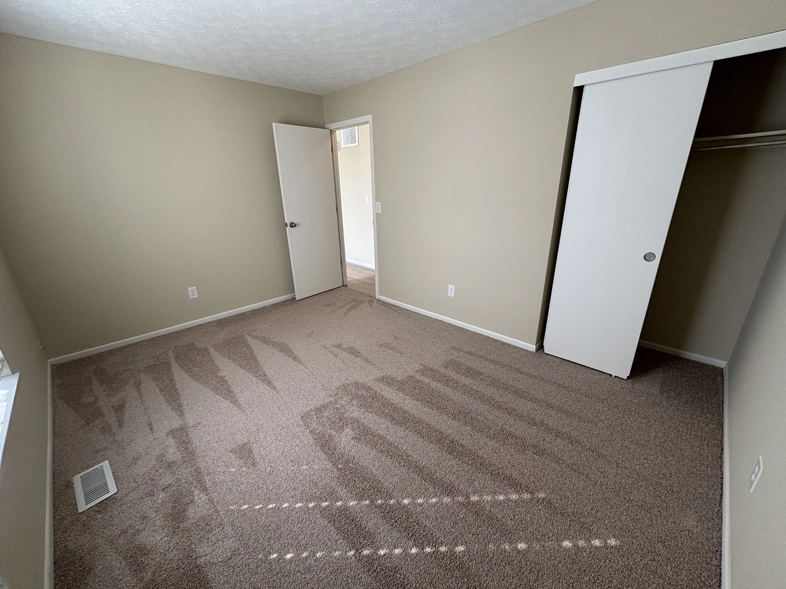 Empty bedroom with beige walls, brown carpet, an open door leading to a hallway, and a closet with a sliding door on the right.