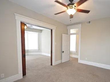 Empty room with beige walls, a ceiling fan, and door leading to a small sunroom with windows.