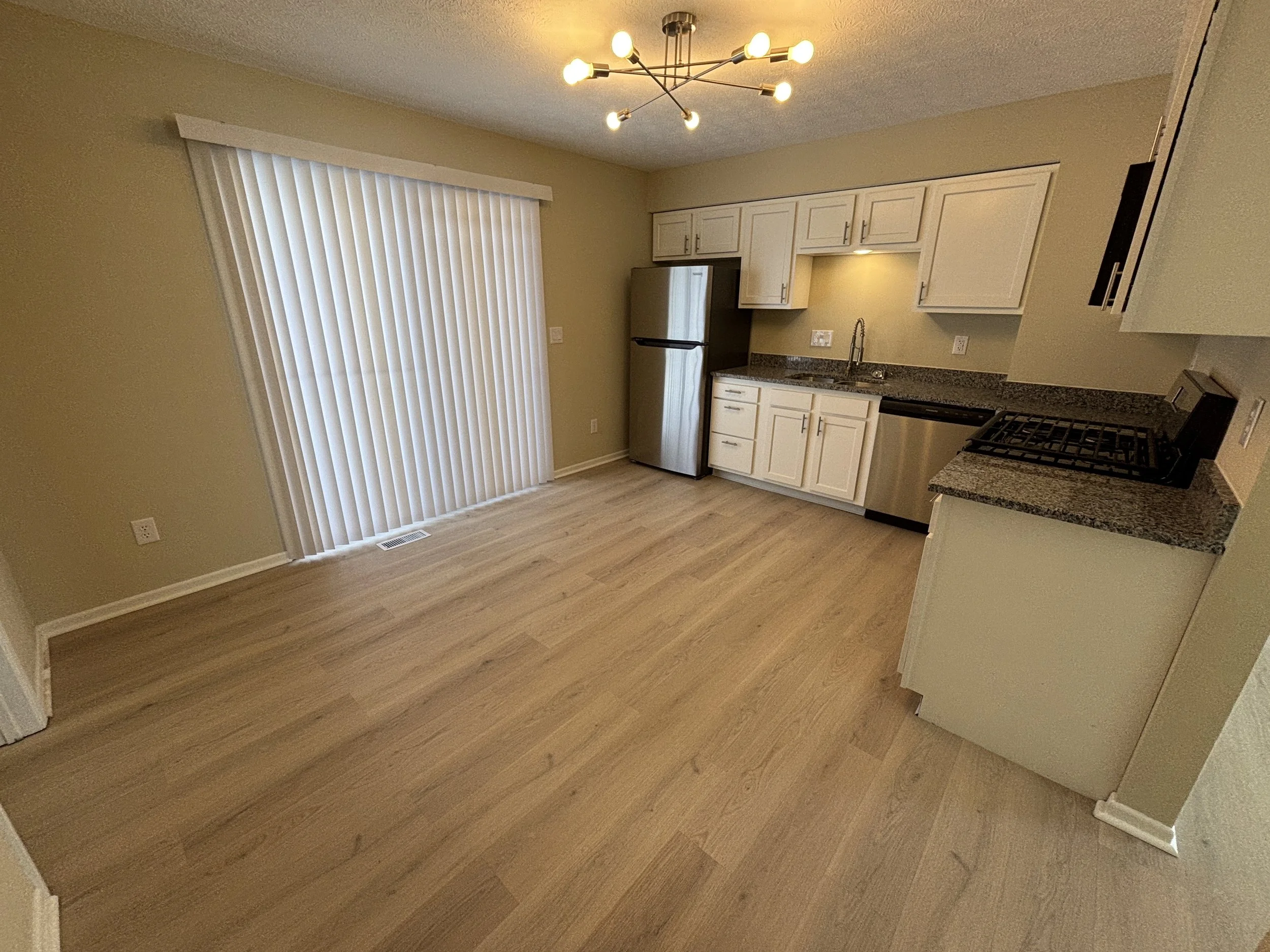 Empty kitchen with light-colored hardwood flooring, white cabinets, stainless steel appliances, a black countertop, and vertical blinds on a sliding glass door, with a modern chandelier overhead.