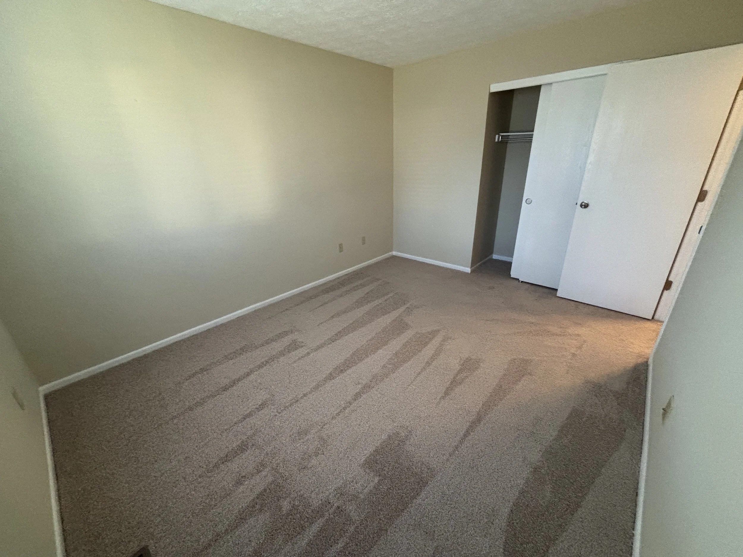Empty bedroom with beige walls, beige carpet with vacuum lines, an open closet with sliding doors, and a partially visible door.