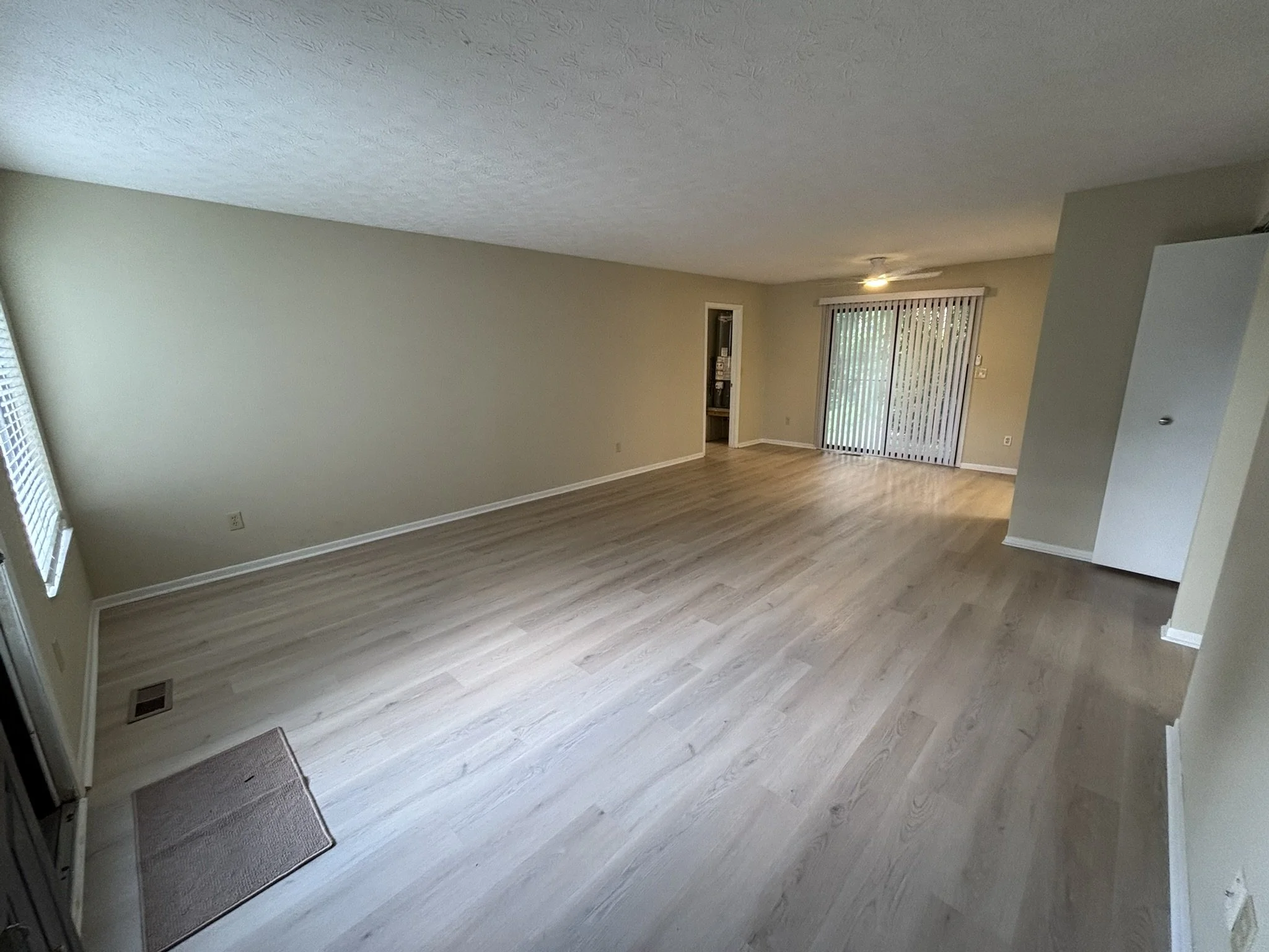 Empty living room with beige walls, light wood flooring, a sliding door with vertical blinds, a window with blinds, and a small opening to another room.