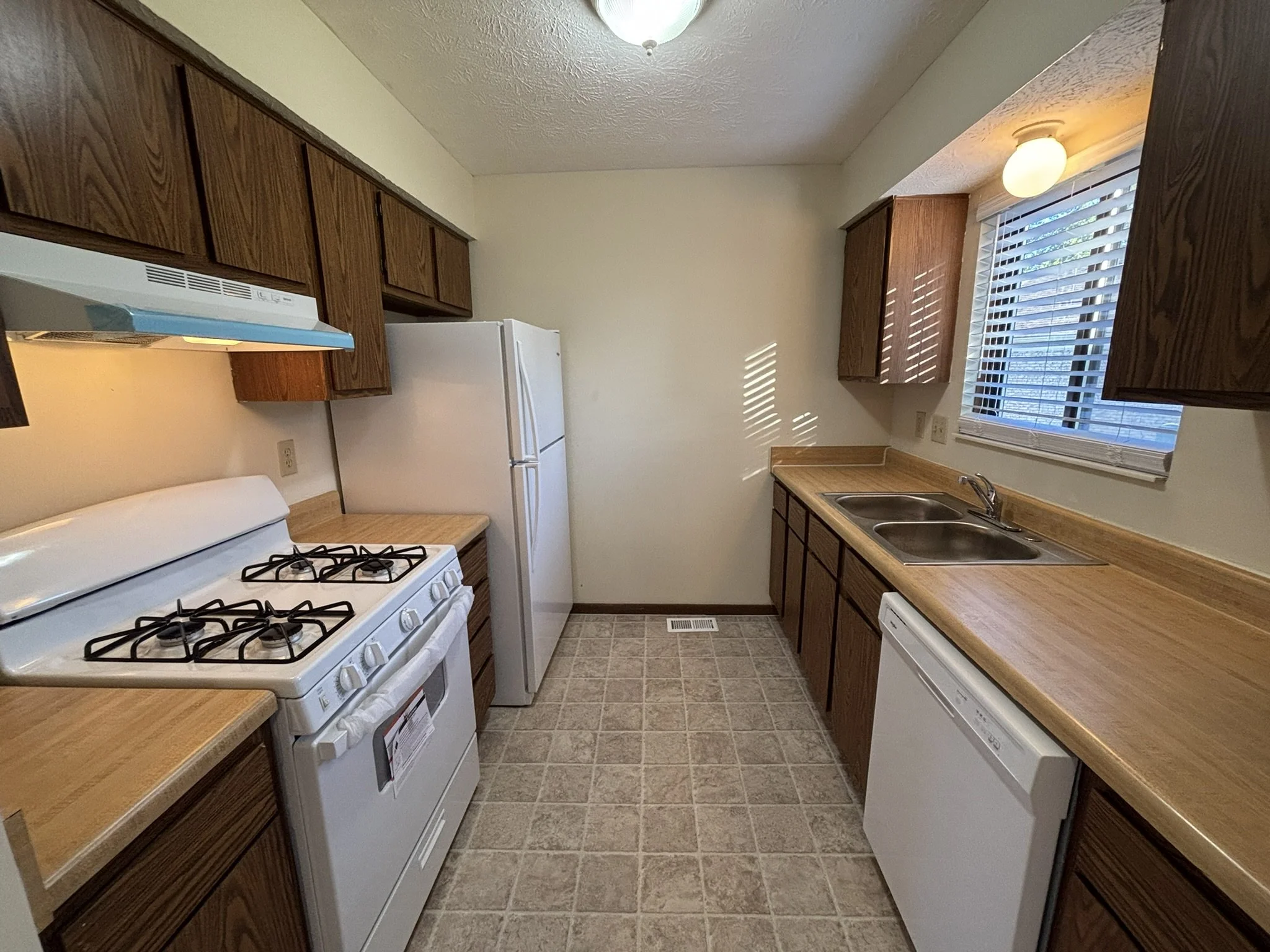 Empty kitchen with wooden cabinets, white appliances including stove, refrigerator, and dishwasher, double sink beneath a window with blinds, tiled floor, and ceiling light
