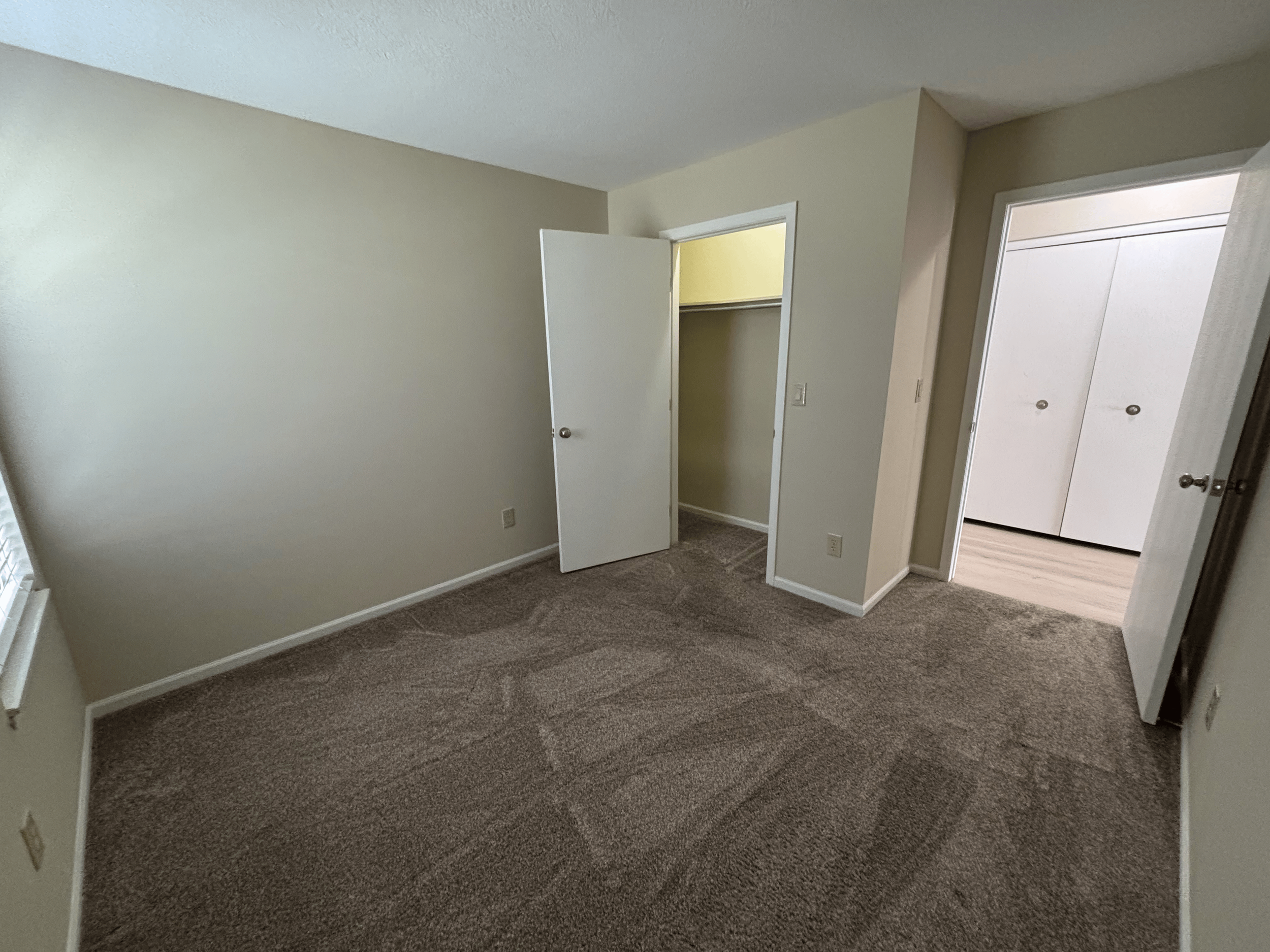 Empty bedroom with beige walls, brown carpet, window on the left, open closet door in the middle, and doorway to another room on the right.