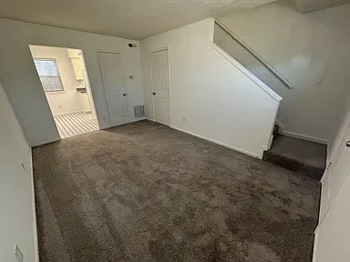Empty living room with beige carpet, white walls, and a staircase leading to an upper floor.