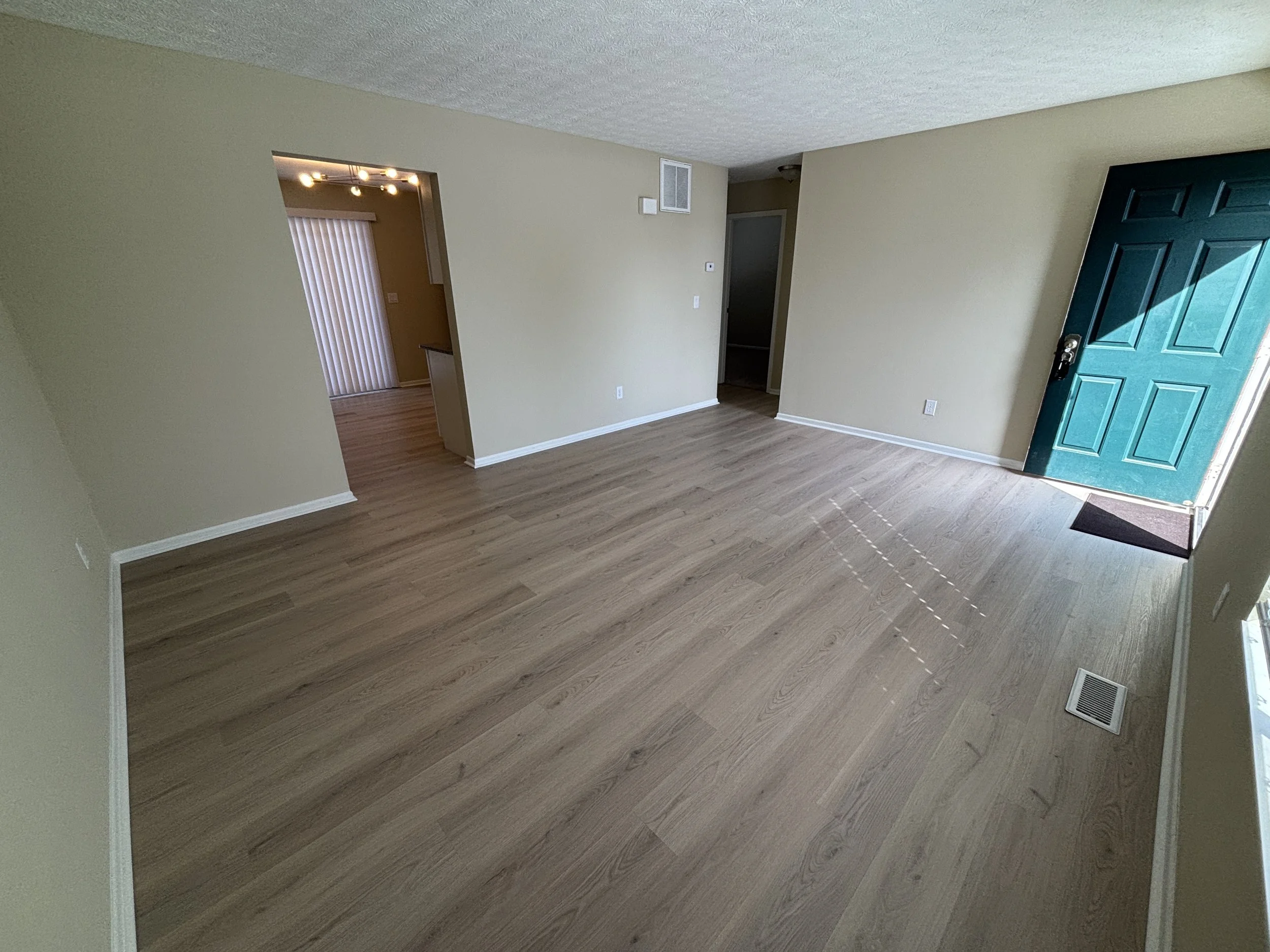 Empty living room with light-colored walls, wood flooring, and a teal front door. Natural light coming through windows, with a view into a dining area with vertical blinds.