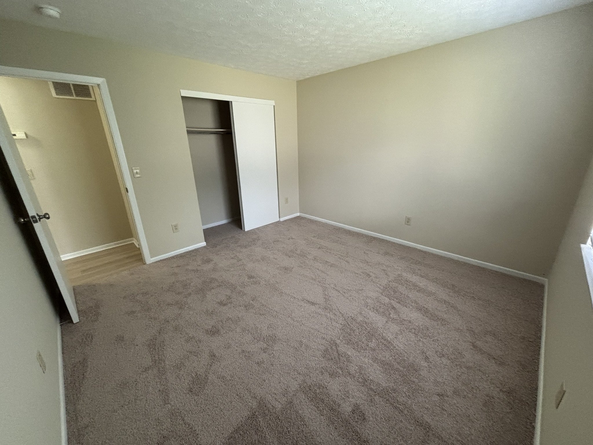 Empty bedroom with beige carpet, beige walls, a closet with a sliding door, and a window on the right side.