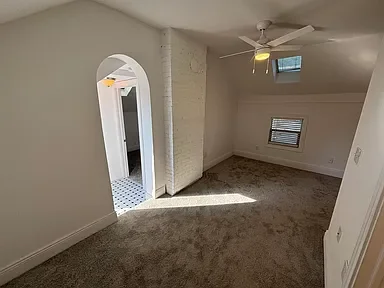 Empty room with beige carpet, two small windows, ceiling fan, and a doorway leading to another room with tiled floor.