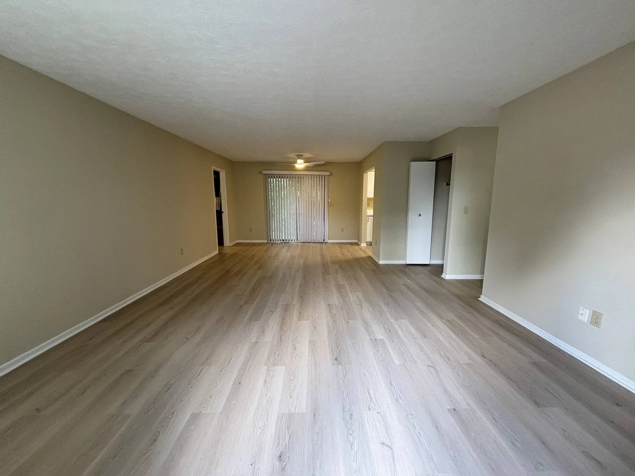 Empty living room with light wood flooring, beige walls, and sliding glass door with vertical blinds leading to patio or balcony.