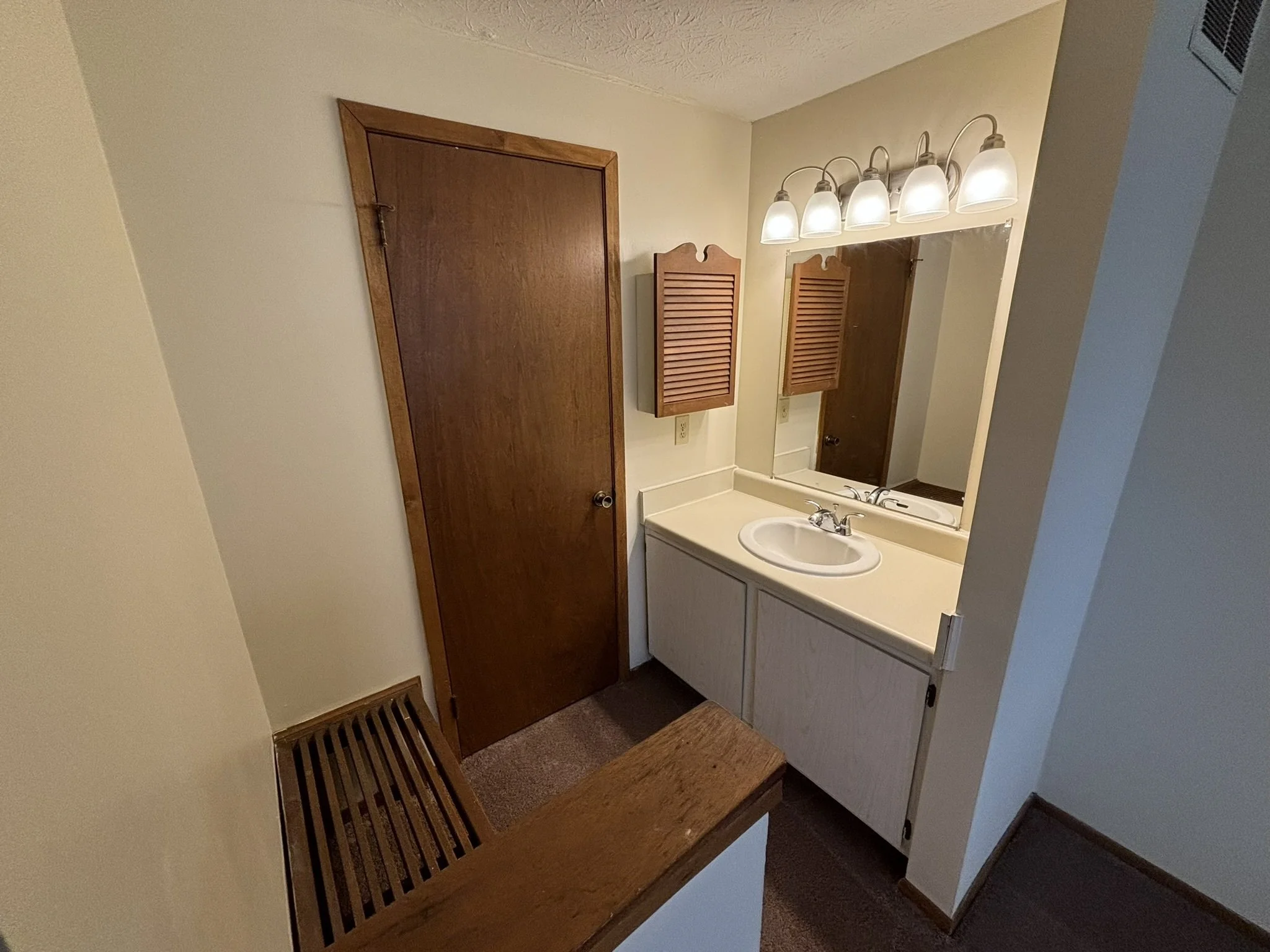 Small bathroom with a single sink and a large mirror. Wooden cabinet doors are above the sink, and there is a row of five light bulbs above the mirror. A wooden bench sits at the entrance.