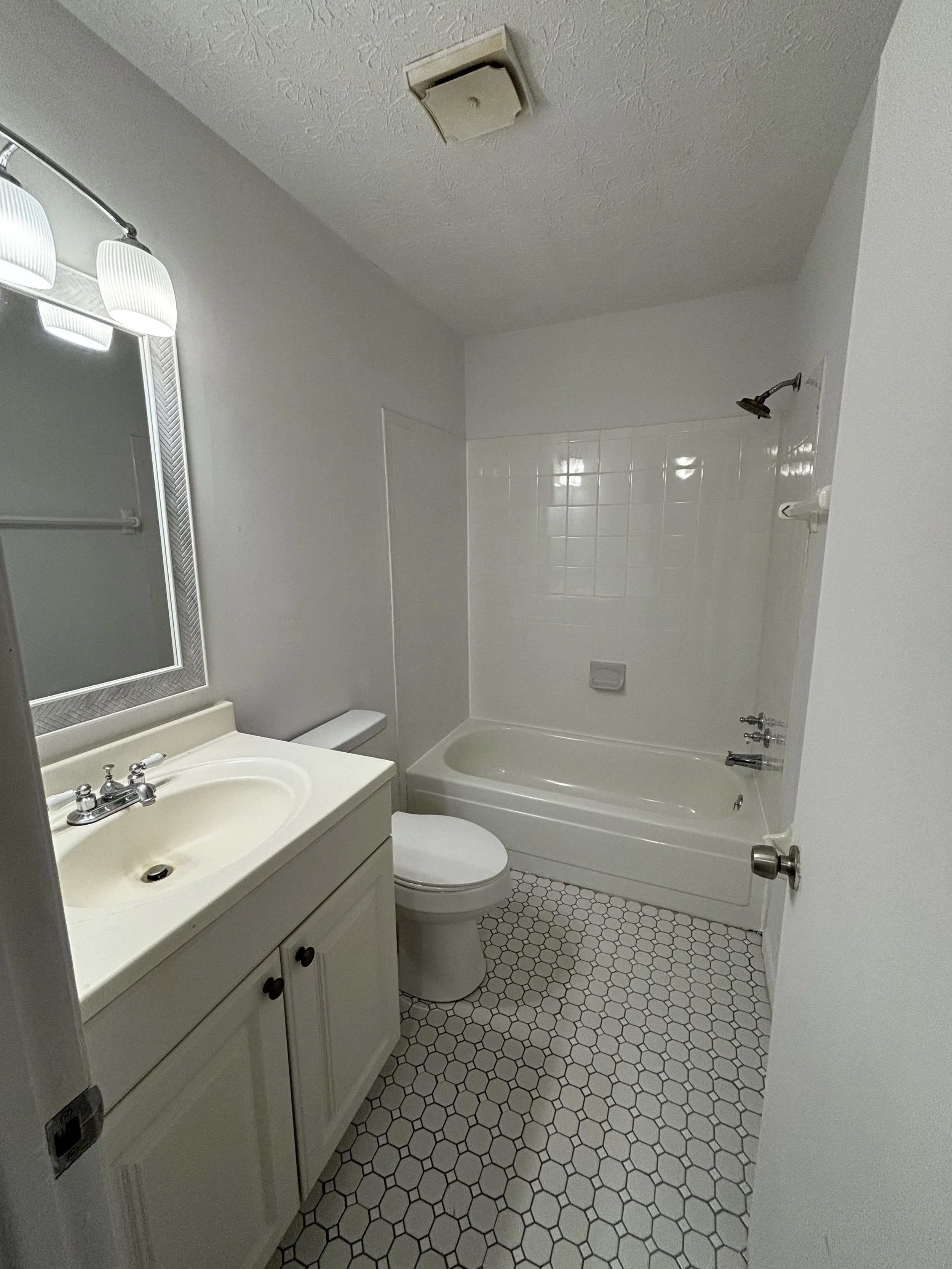 Small bathroom with white hexagonal tiled floor, white vanity with a sink, white toilet, bathtub with white tile surround, mirror with lights, ceiling vent, and walls painted white.