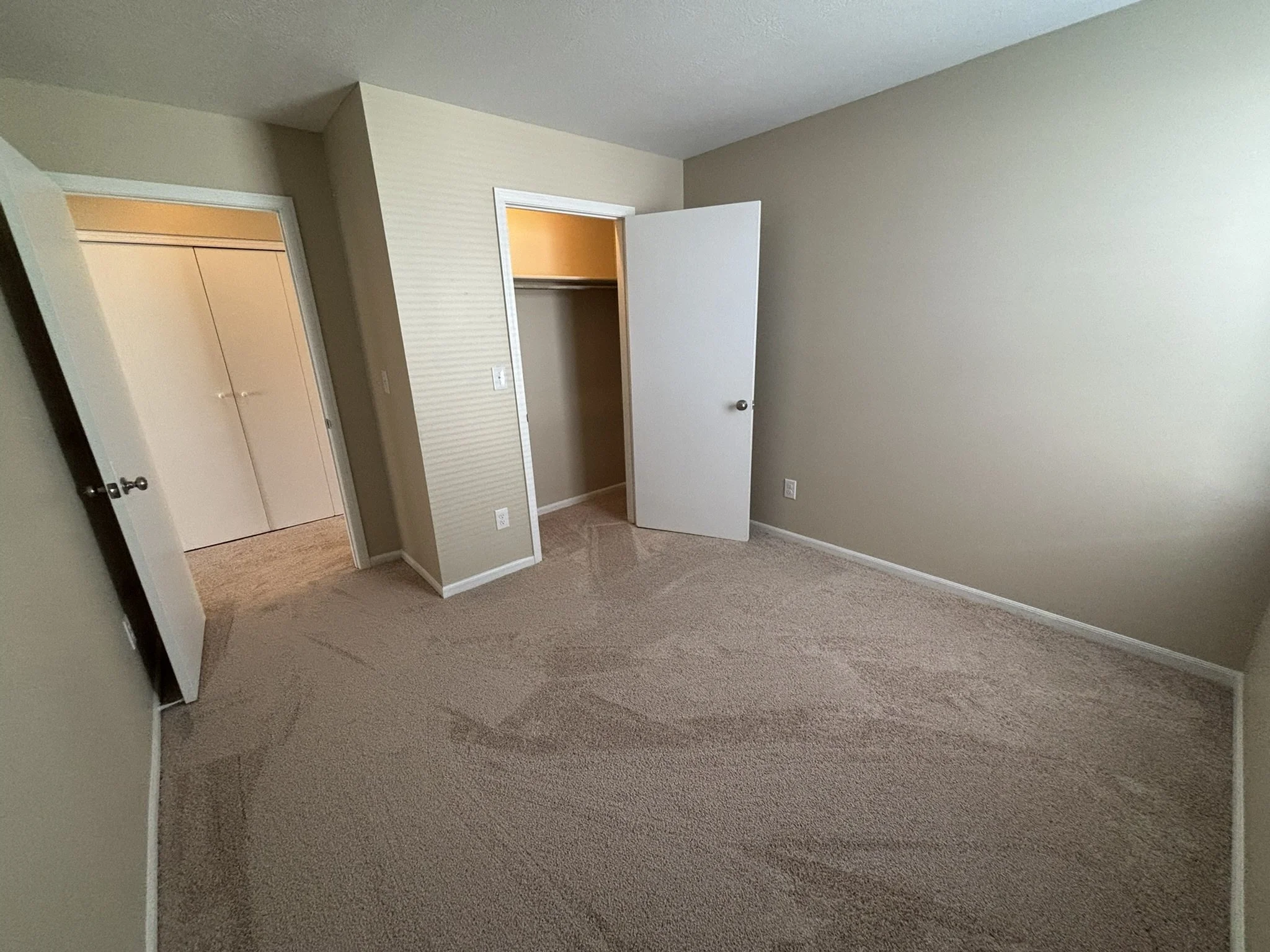 Empty bedroom with beige carpet, off-white walls, open closet door, and closed closet with double doors.