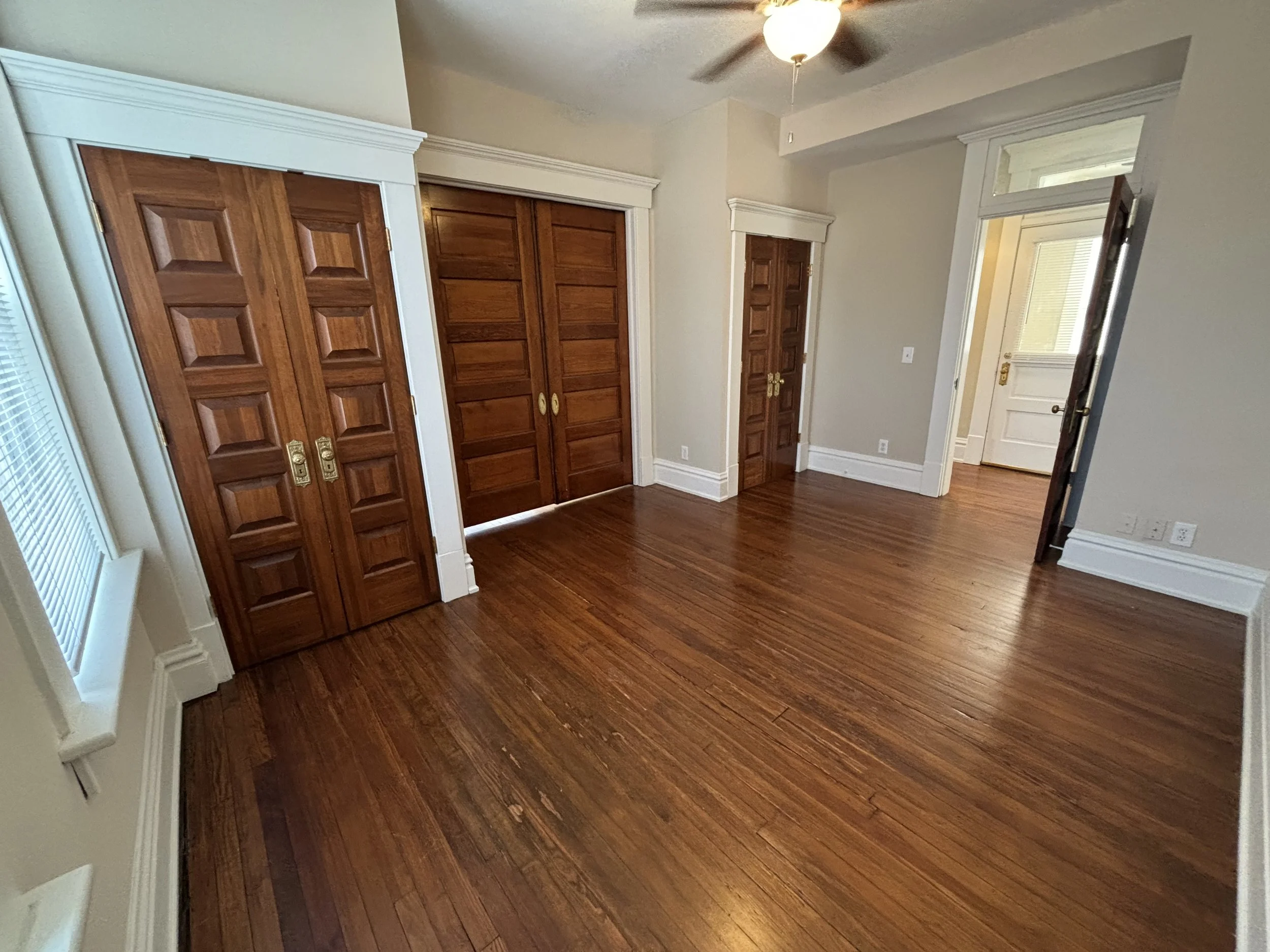 Empty room with wooden floors, a ceiling fan, and multiple closed wooden closet doors. There is a window with blinds on the left side.