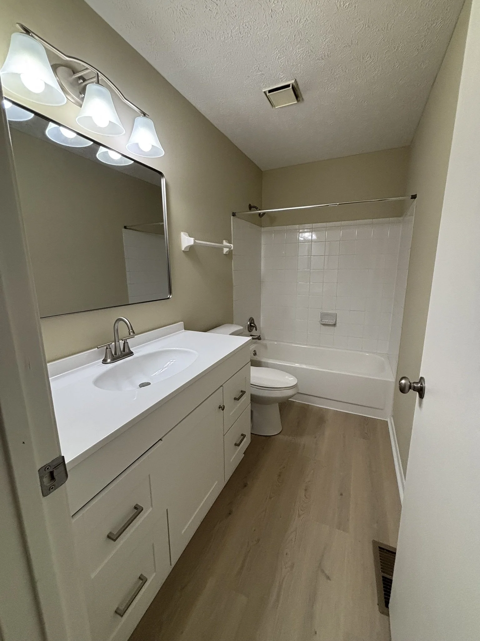 A small bathroom with a white vanity sink, a mirror above it, a toilet, and a bathtub with a shower curtain rod. The walls are painted beige, and the floor is made of light wood or laminate. There is a ceiling vent and a light fixture with multiple b