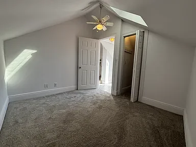 Empty room with beige carpet, white walls, two open doors, ceiling fan, and natural light from a window.