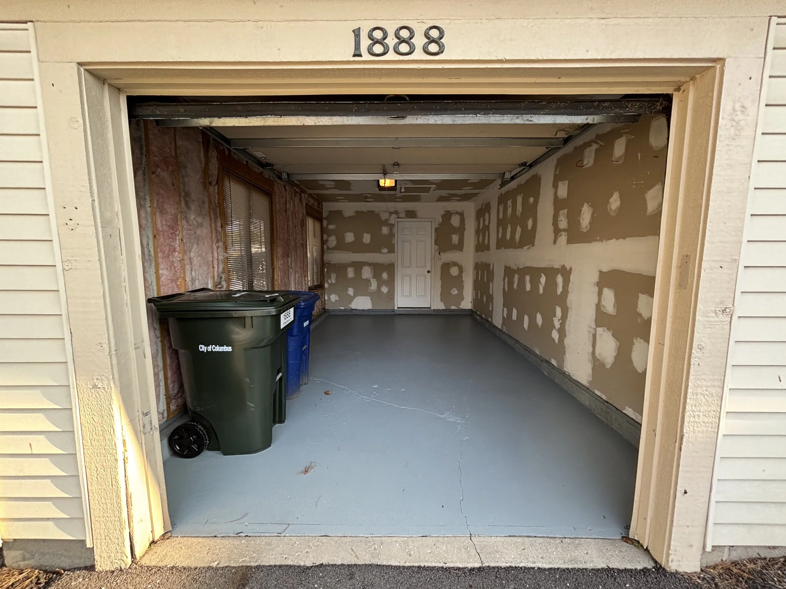 Empty garage with unfinished drywall, a white door at the back, and three trash bins outside labeled City of Columbus, with the number 1888 above the garage door