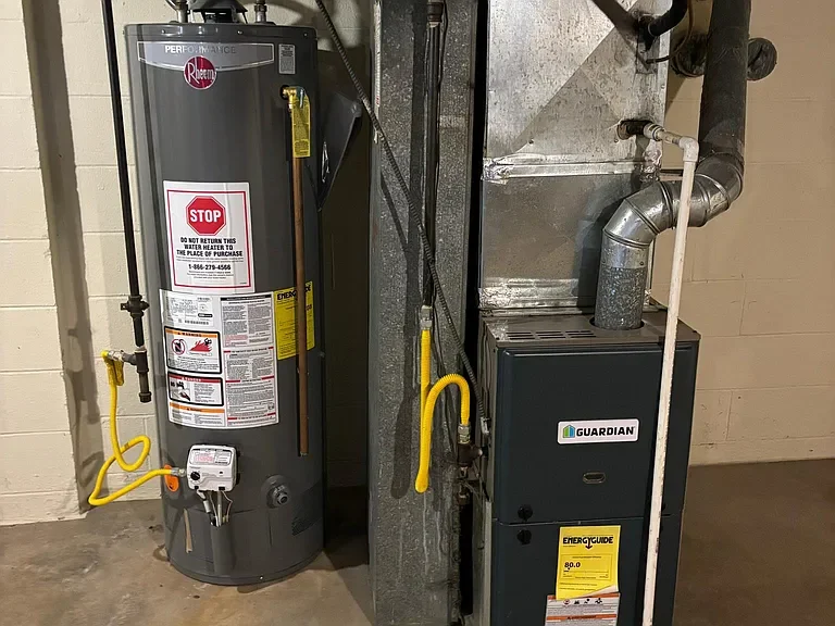 A water heater with various labels and a yellow gas line, standing next to a furnace in a utility room with a concrete floor and beige cinder block wall.