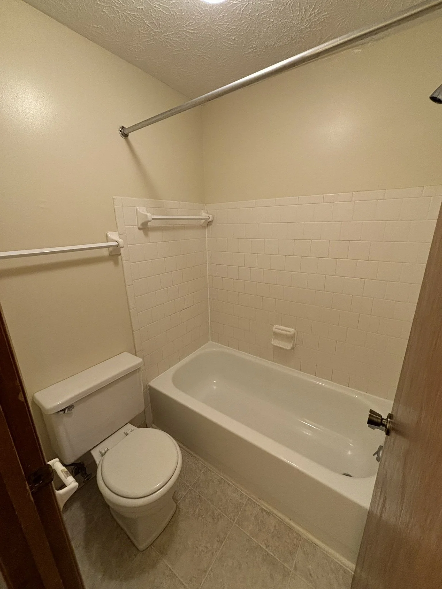 Small bathroom with a white toilet and a bathtub, beige tiled wall, and a beige painted wall, with two towel bars and a shower head.