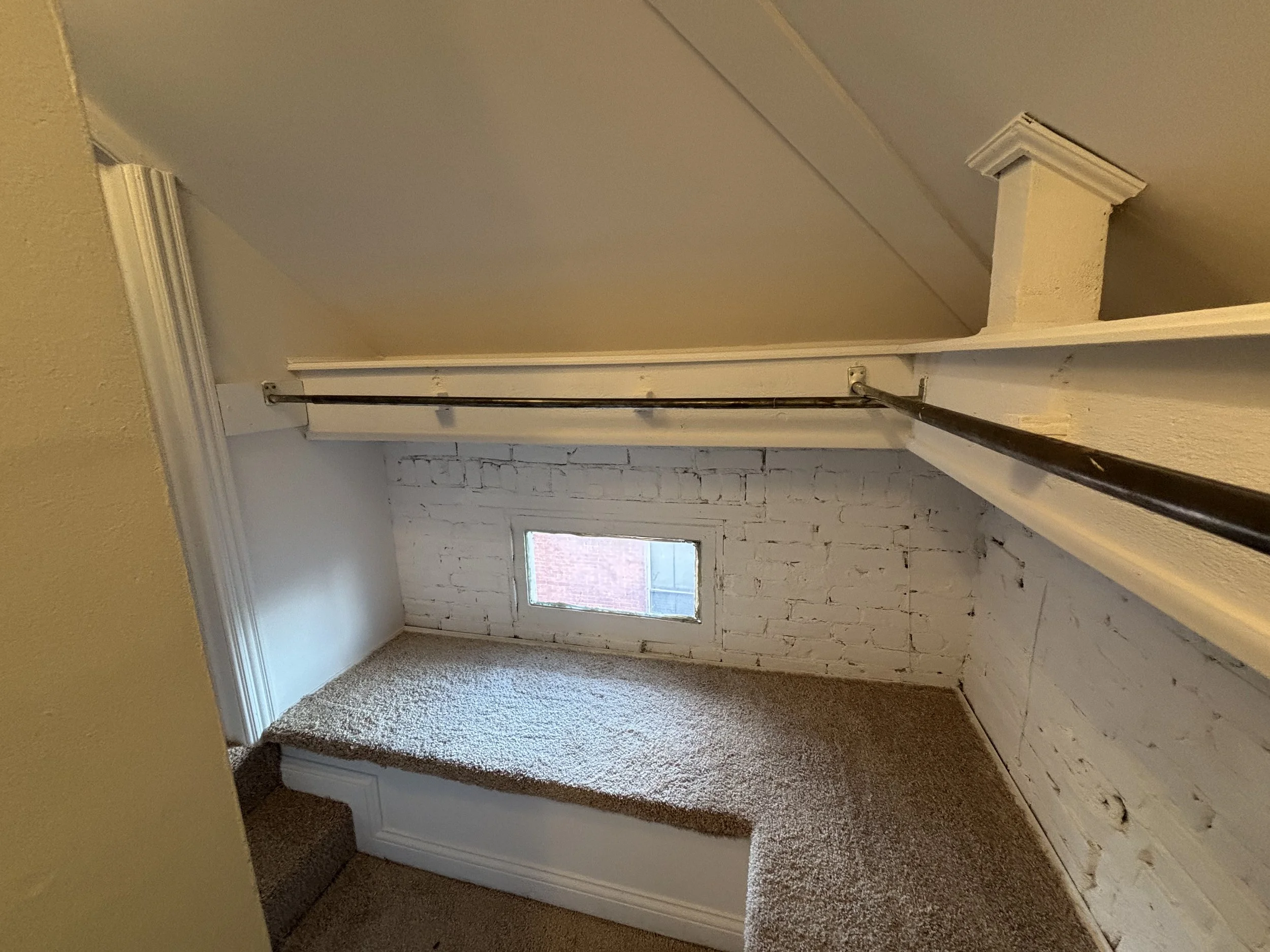 Small loft space with white brick wall, window, beige carpet, and a black rod for hanging clothes.