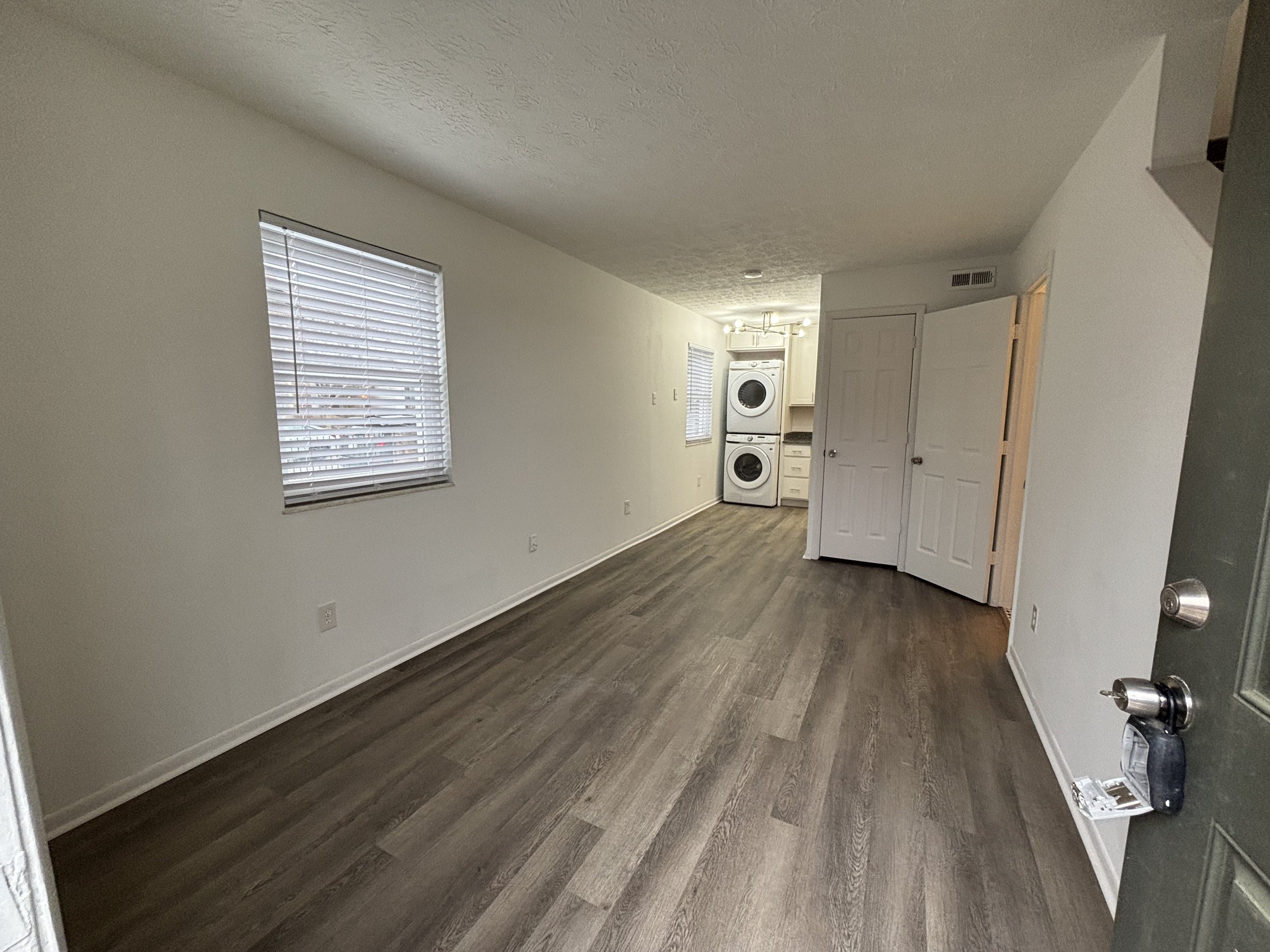 Empty living space with wooden flooring, white walls, two windows with blinds, and a laundry area with stacked washer and dryer in the background.